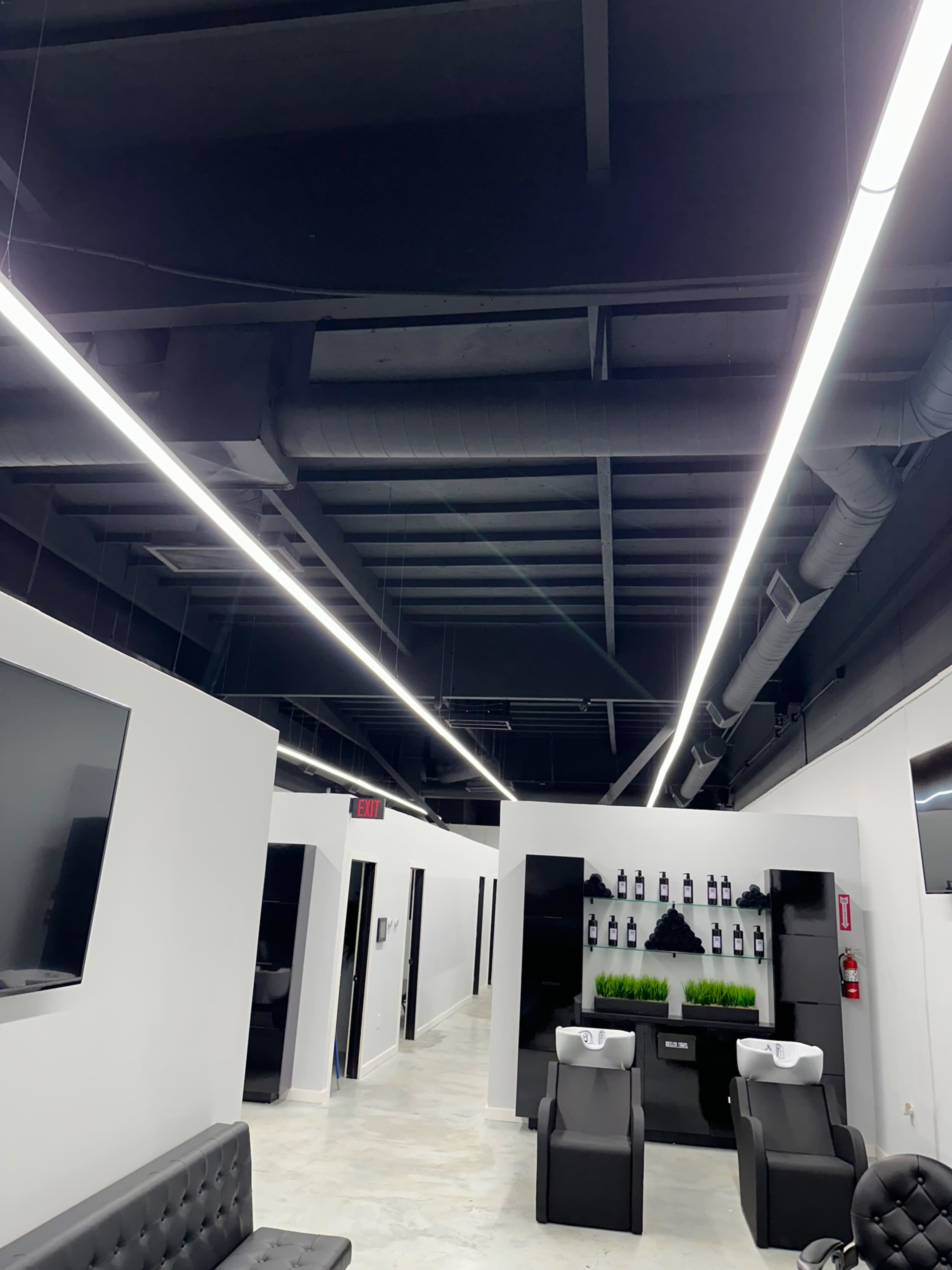 The image shows a modern salon interior with long overhead lights, black chairs, and a clean, minimalistic design.