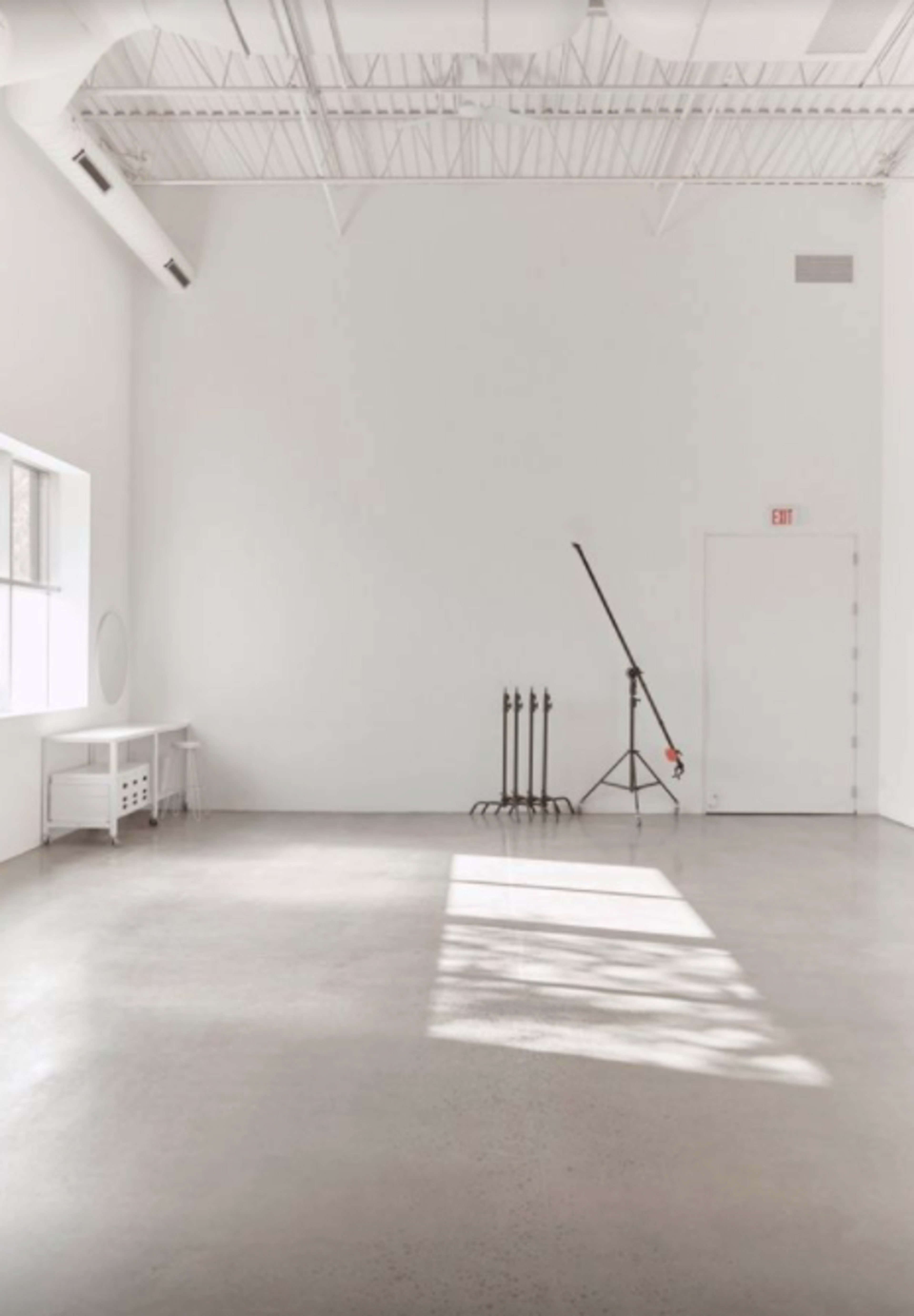 A minimalist, empty room with a concrete floor, a large window, and a lighting stand positioned against the wall.