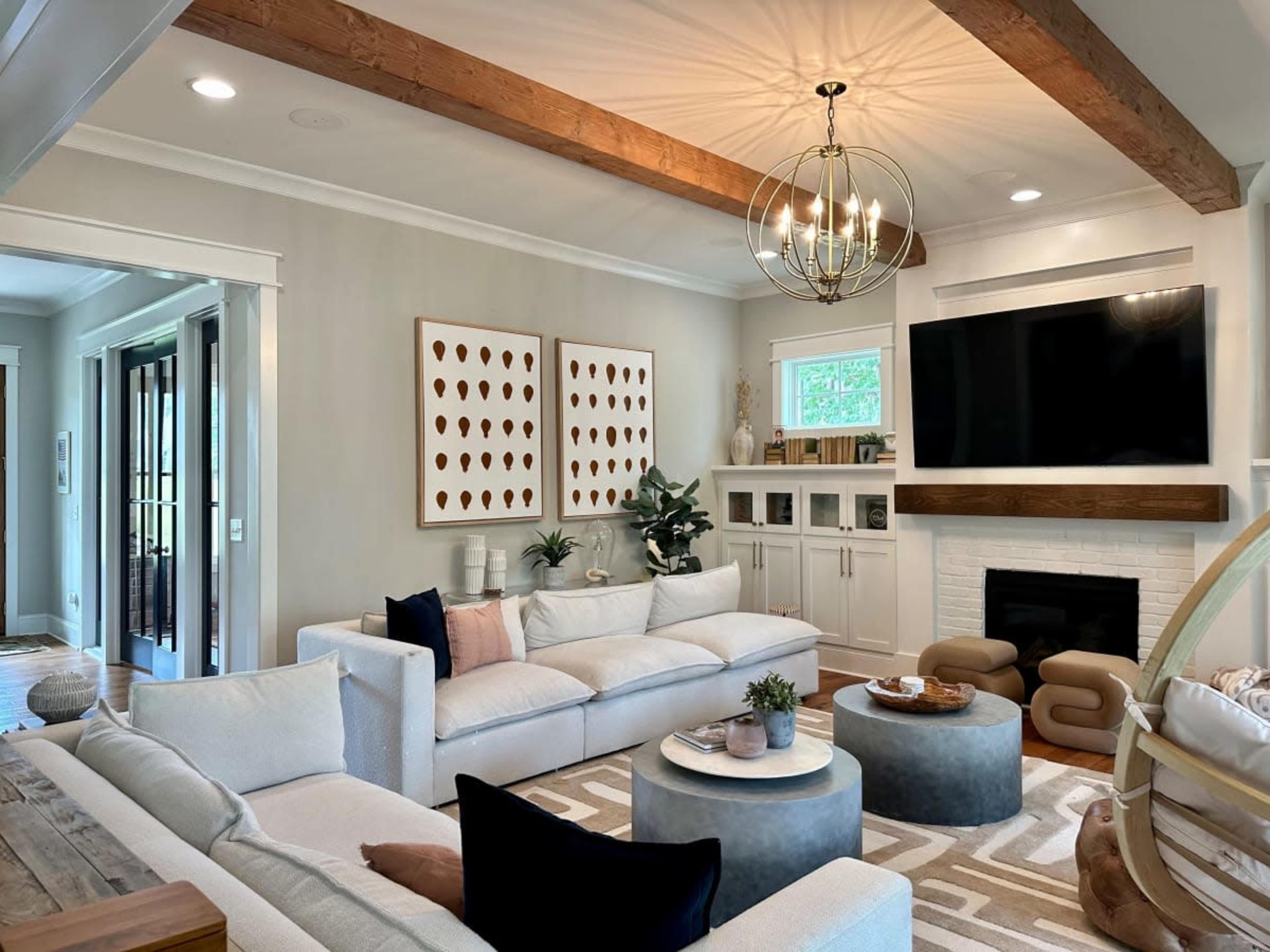 A modern living room features white furniture, a large TV mounted above a fireplace, and decorative art on the walls, with wooden beams and a chic light fixture overhead.