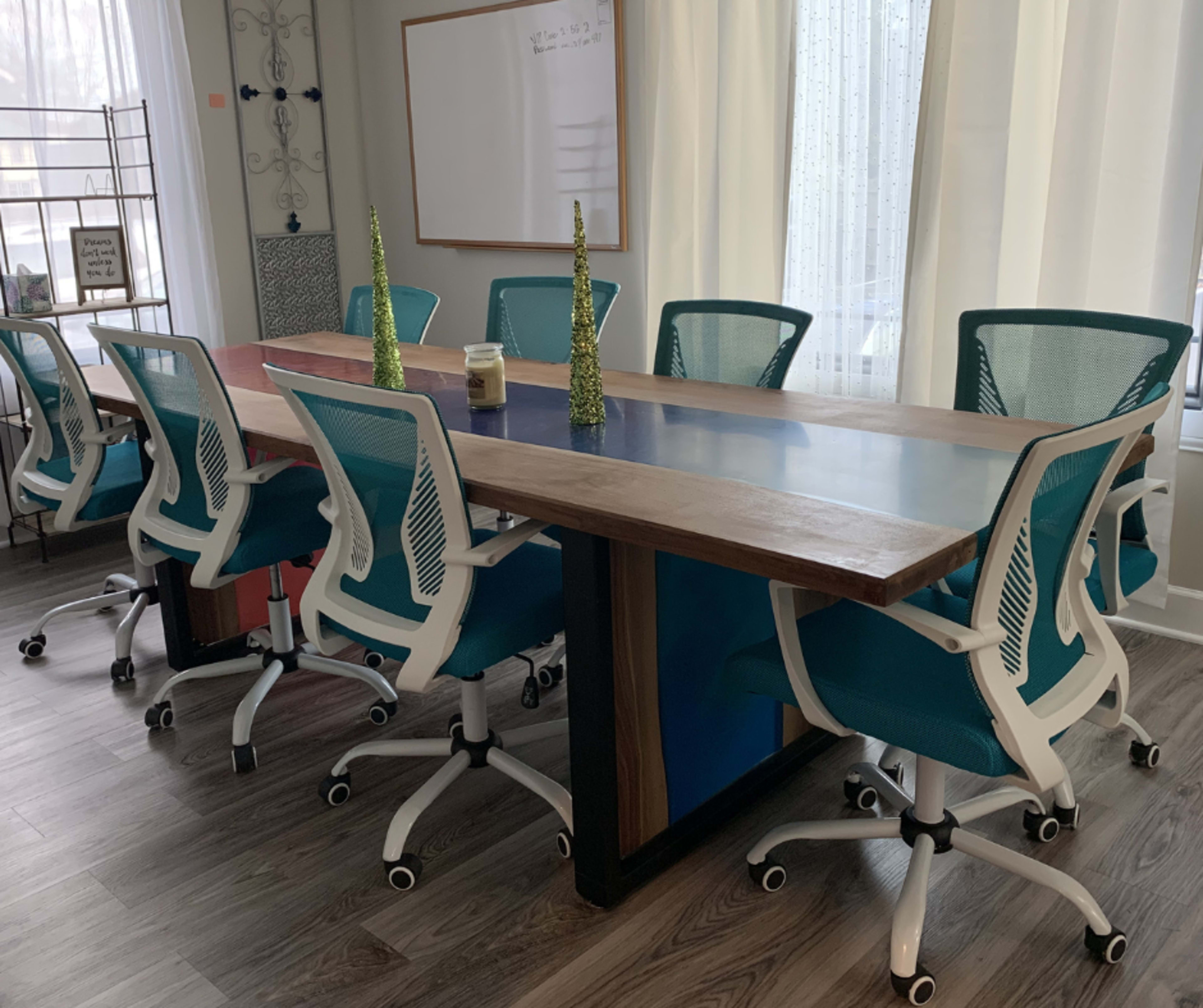 A modern conference room features a long wooden table with a glass center and several teal office chairs arranged around it.