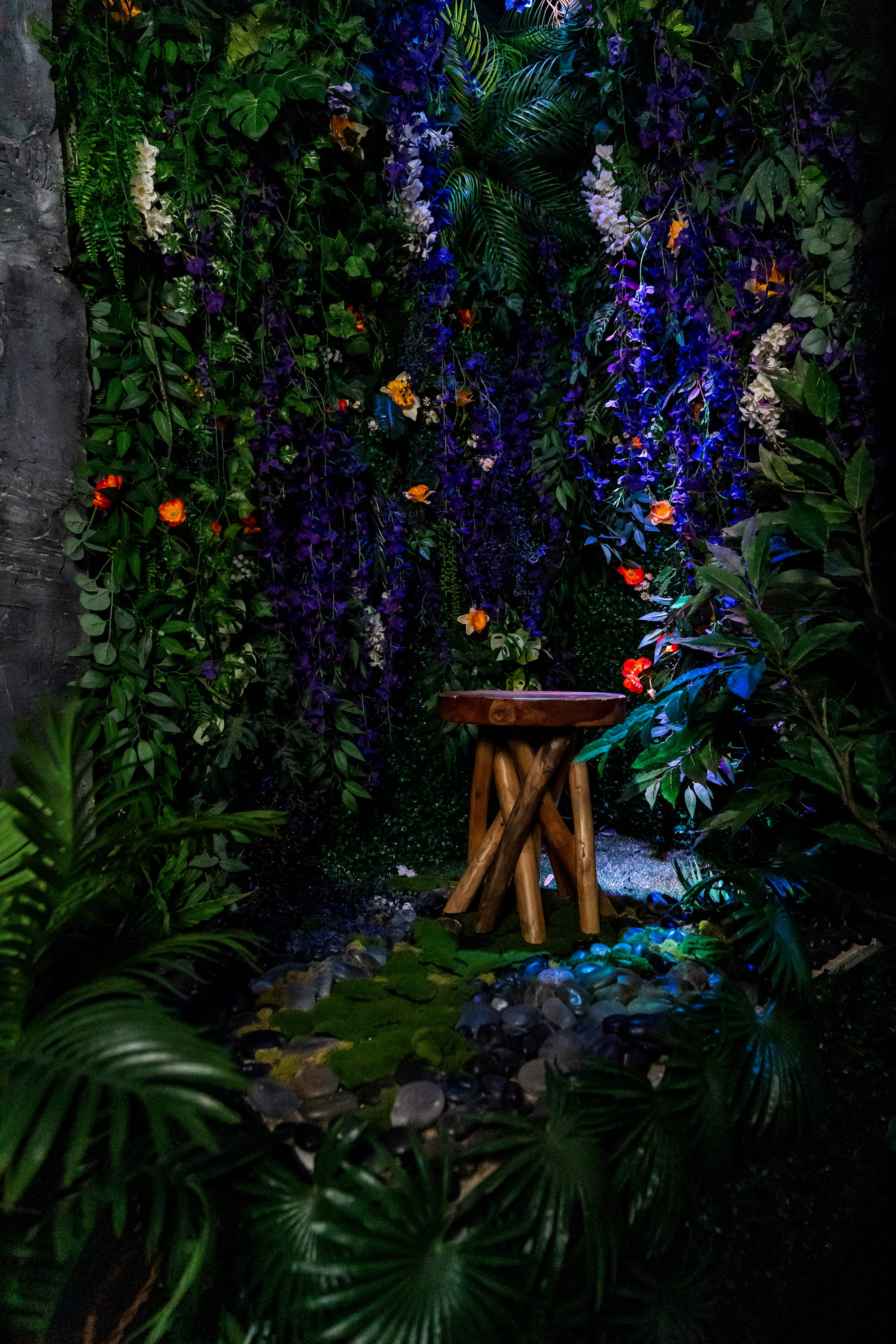 A wooden stool stands amidst a lush backdrop of vibrant flowers and leafy greenery in a serene garden setting.