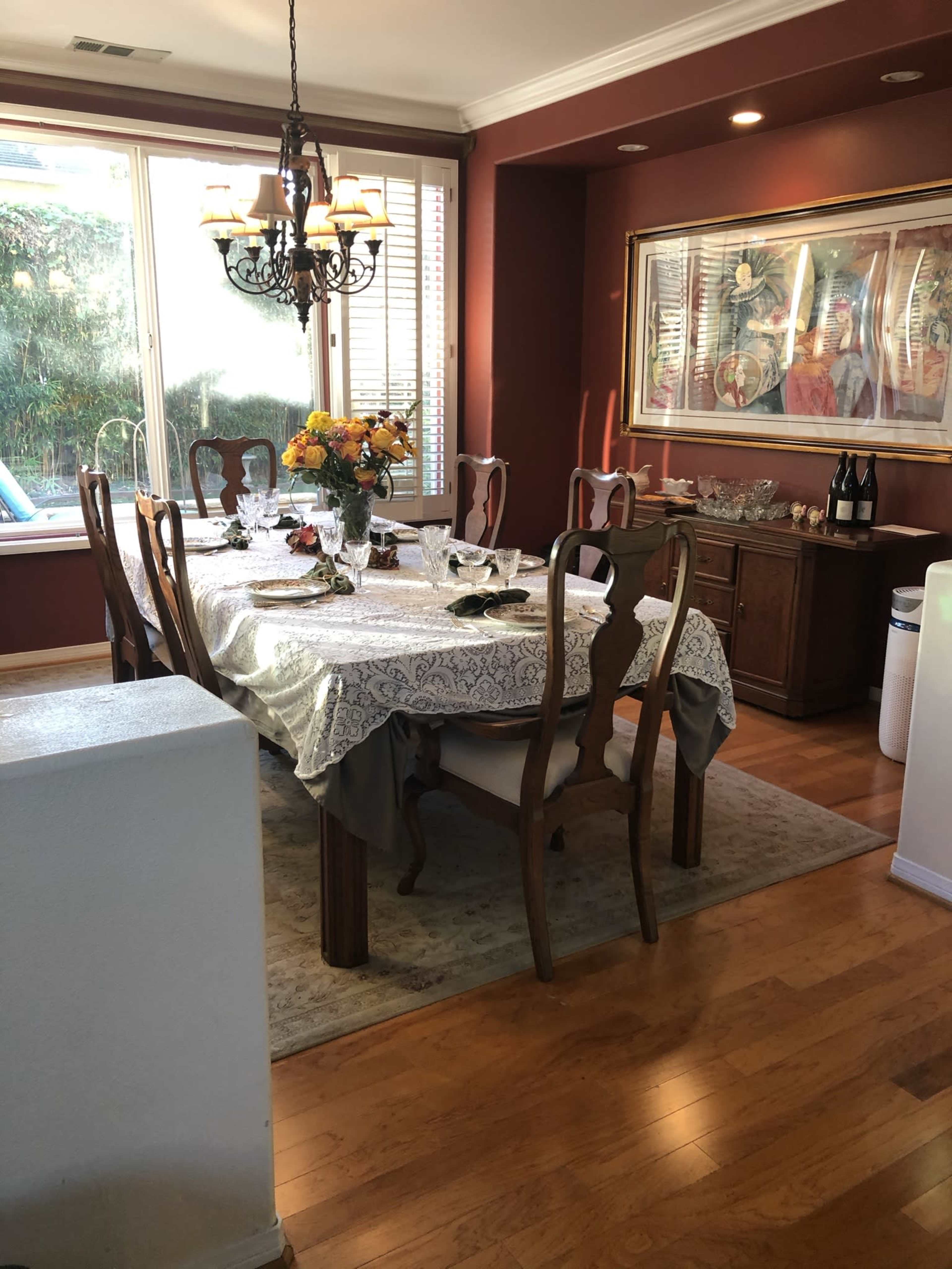 A dining room features a wooden table set with dishes and a floral centerpiece, surrounded by chairs and illuminated by a chandelier.