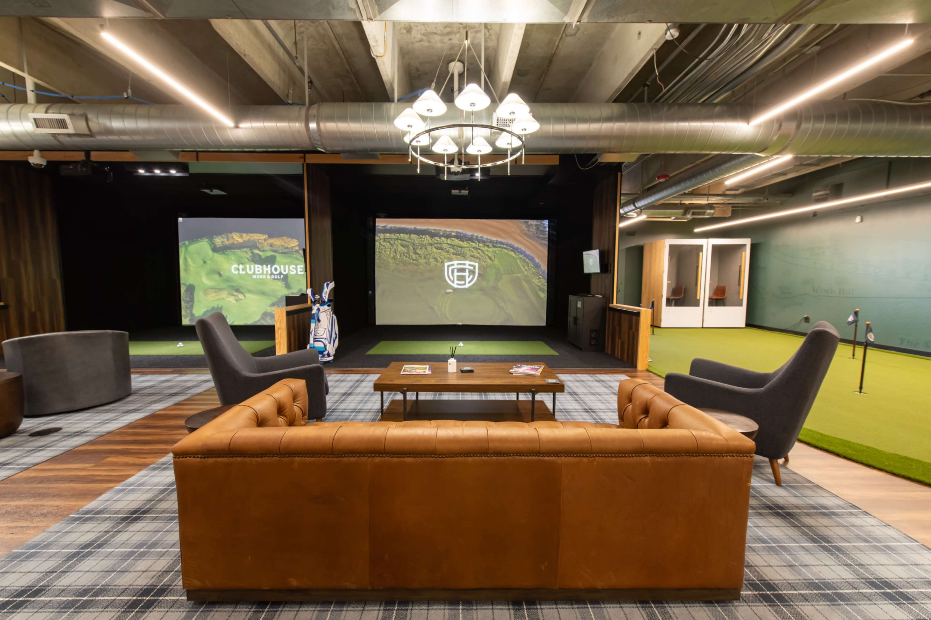 The image shows a modern lounge area designed for golfing, featuring a brown leather sofa, a coffee table, and two chairs beside a putting green and projection screens displaying golfing content.