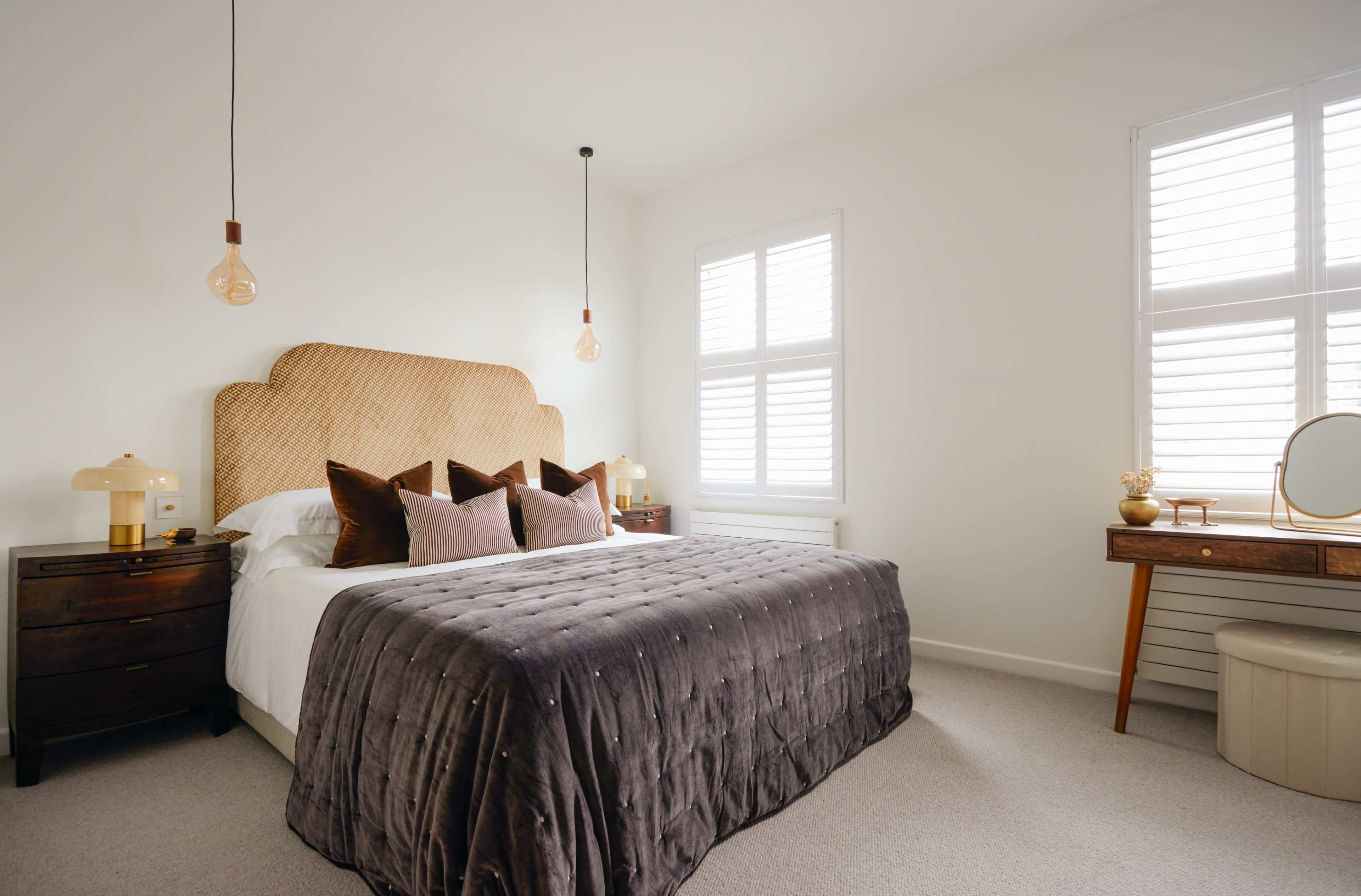 A neatly arranged bedroom featuring a large bed with a quilted cover, two pendant lights, and wooden furniture, including a dresser and a circular vanity.