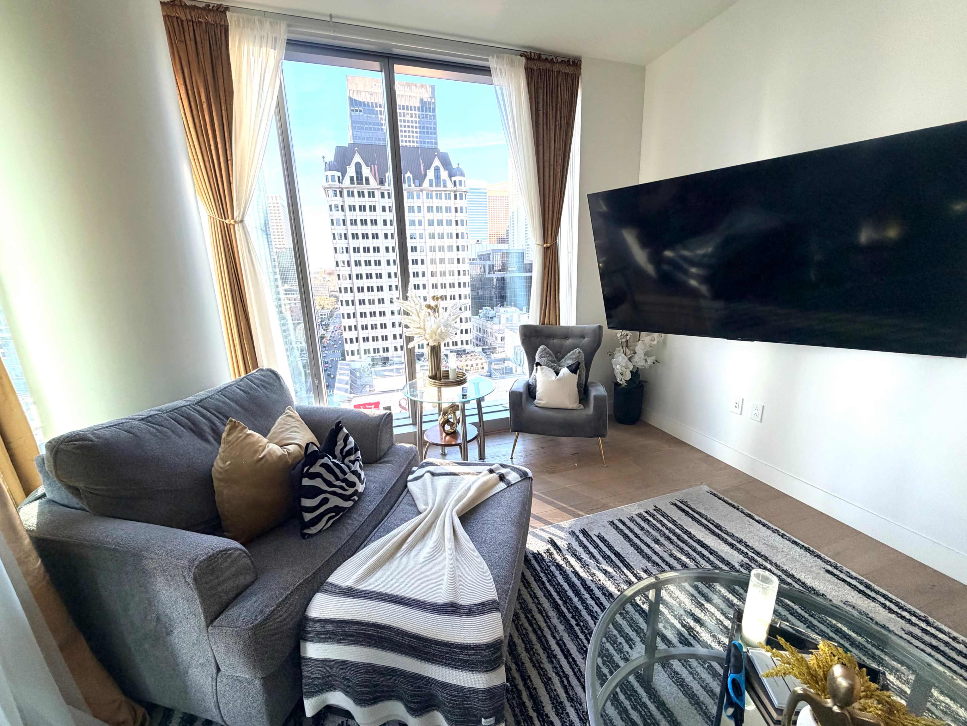The image shows a modern living room with a gray sofa, a glass coffee table, and a large television mounted on the wall, with a view of city buildings through a large window.
