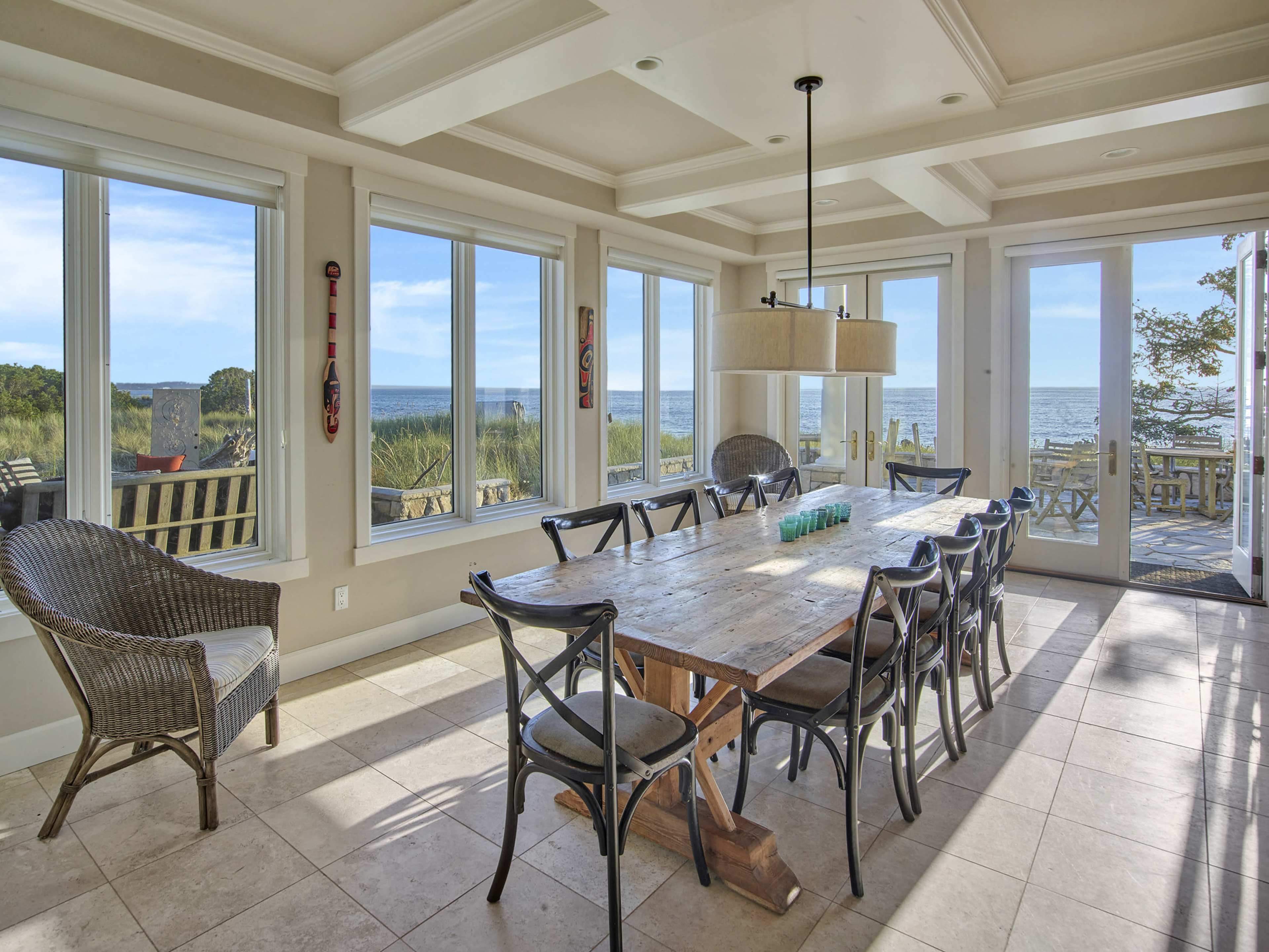 A spacious dining area features a large wooden table surrounded by black chairs, with large windows offering a view of the ocean.