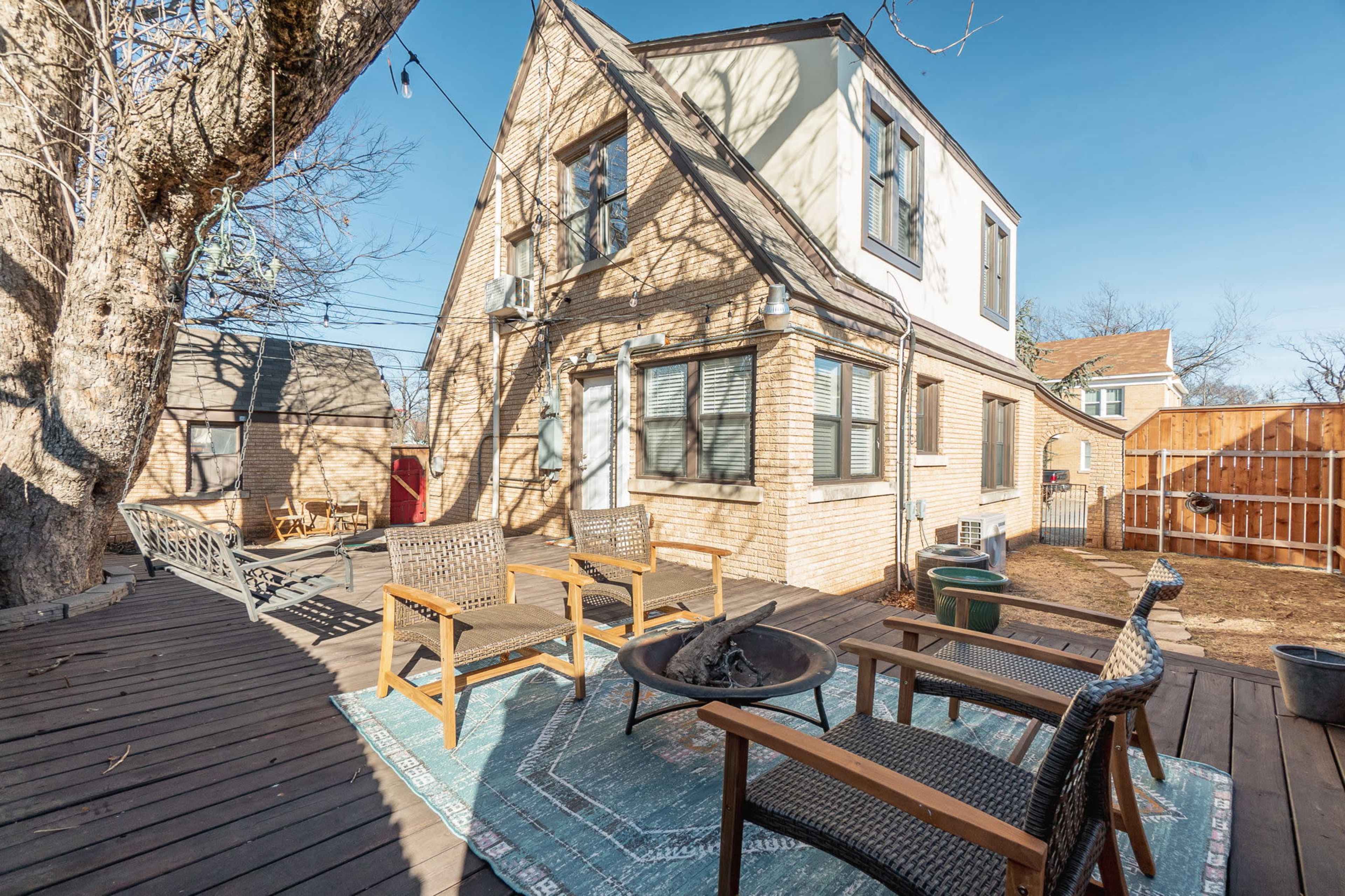 A backyard with a wooden deck featuring seating chairs around a fire pit, adjacent to a two-story house and a wooden fence.