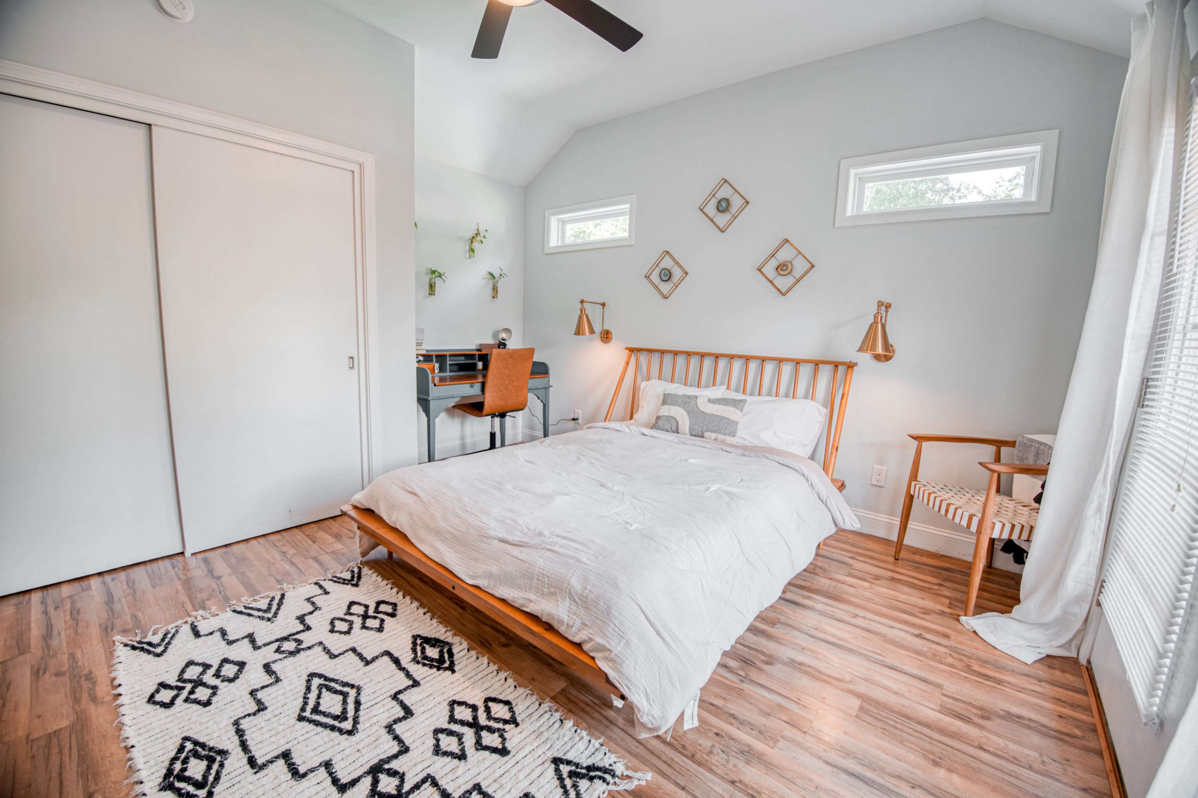 A modern bedroom with a wooden bed, patterned rug, and a small workspace featuring a desk and chair under bright windows.