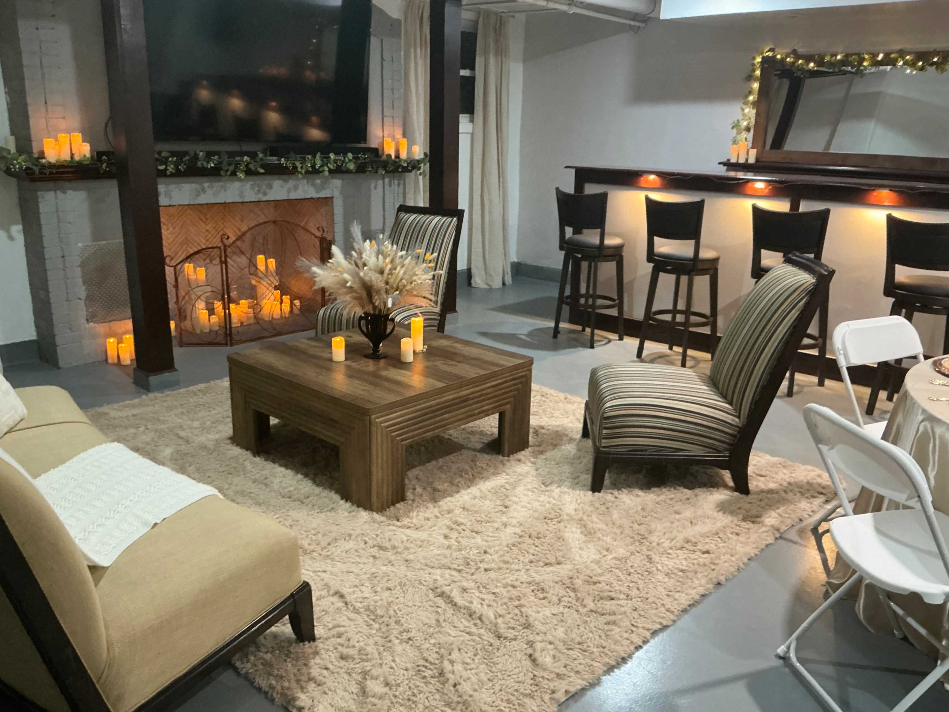 The image shows a cozy living room with a fireplace, a coffee table, two chairs, a bar area, and decorative candles arranged on a soft rug.