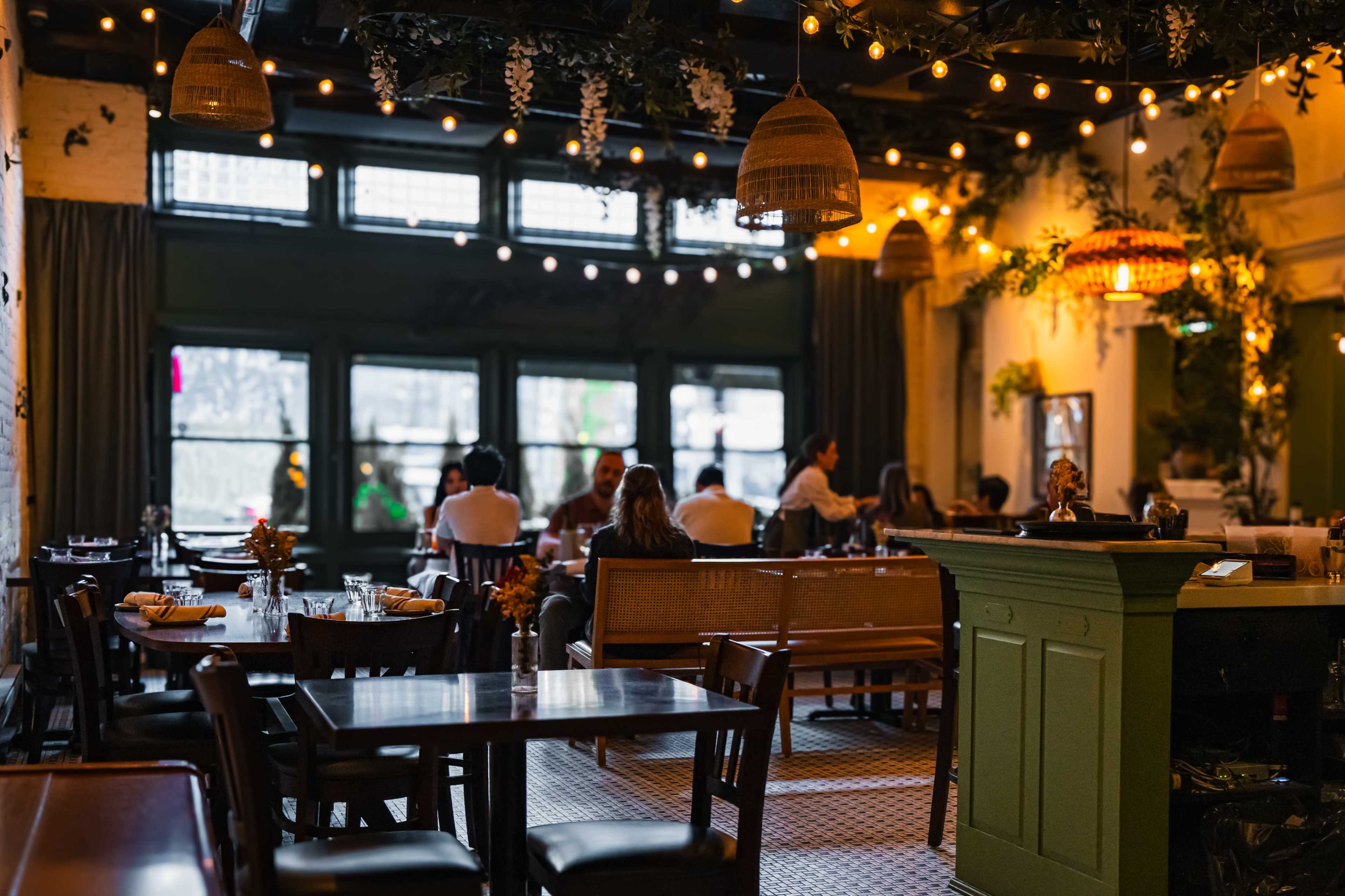 The image shows a restaurant interior with wooden tables, string lights, and greenery, filled with patrons seated at various tables.