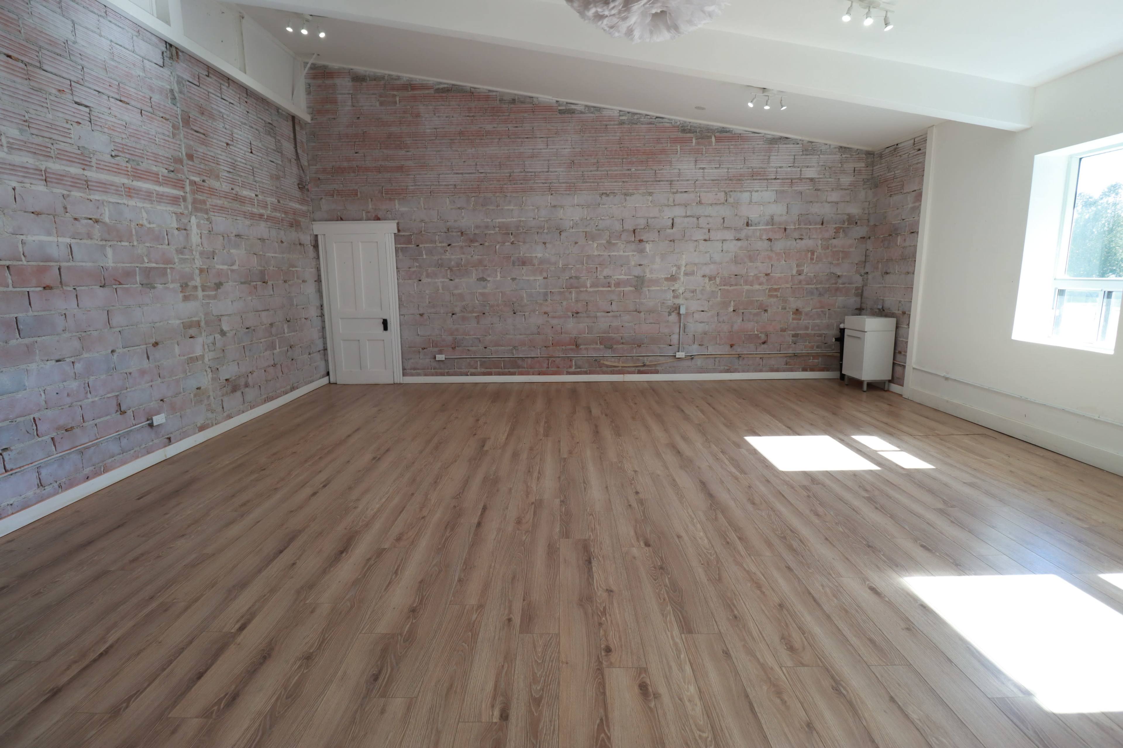 A spacious room with wooden flooring, exposed brick walls, a white door, and a small cabinet in the corner.