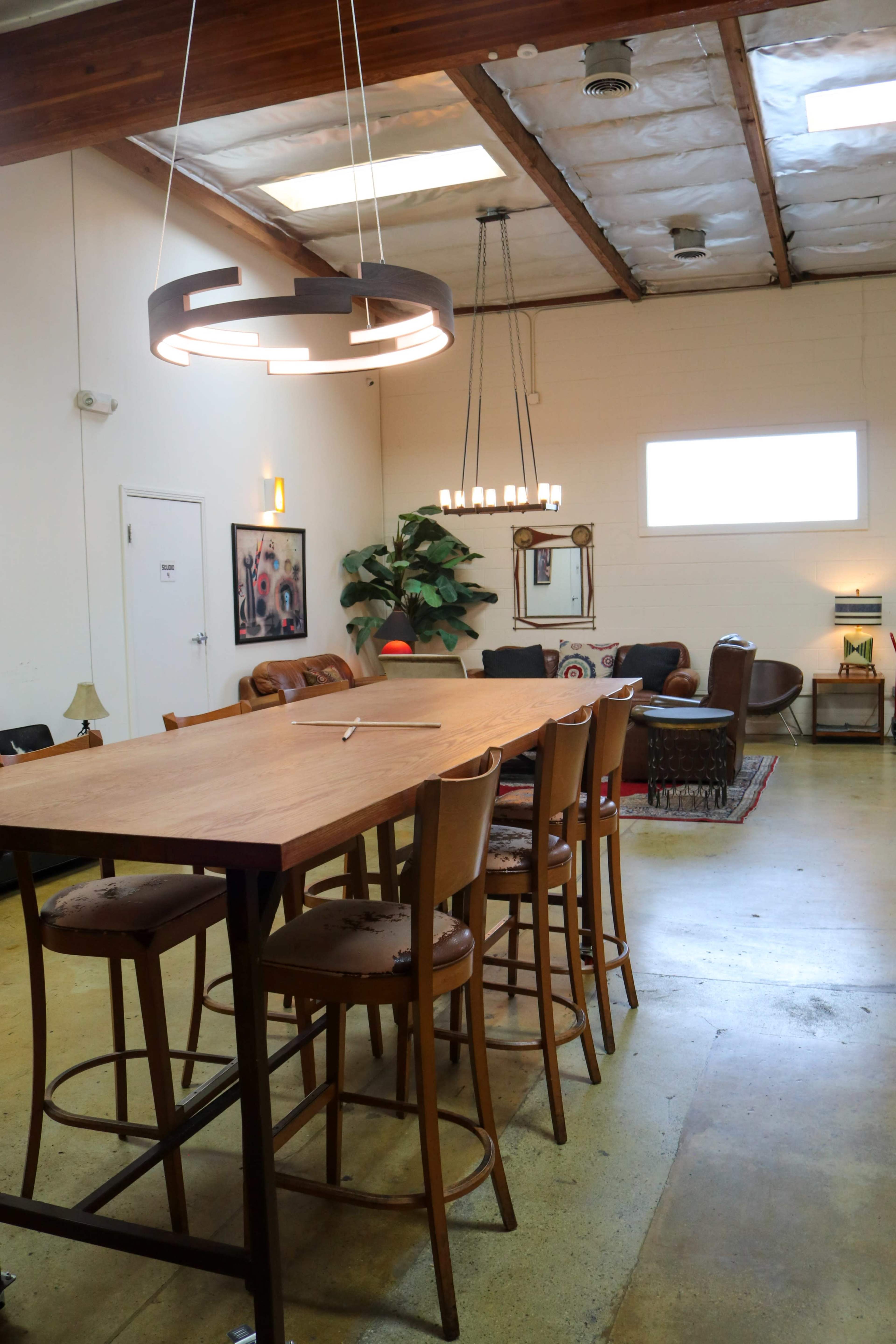 The image shows a spacious room with a wooden dining table surrounded by tall chairs, illuminated by modern light fixtures, and featuring a cozy seating area.