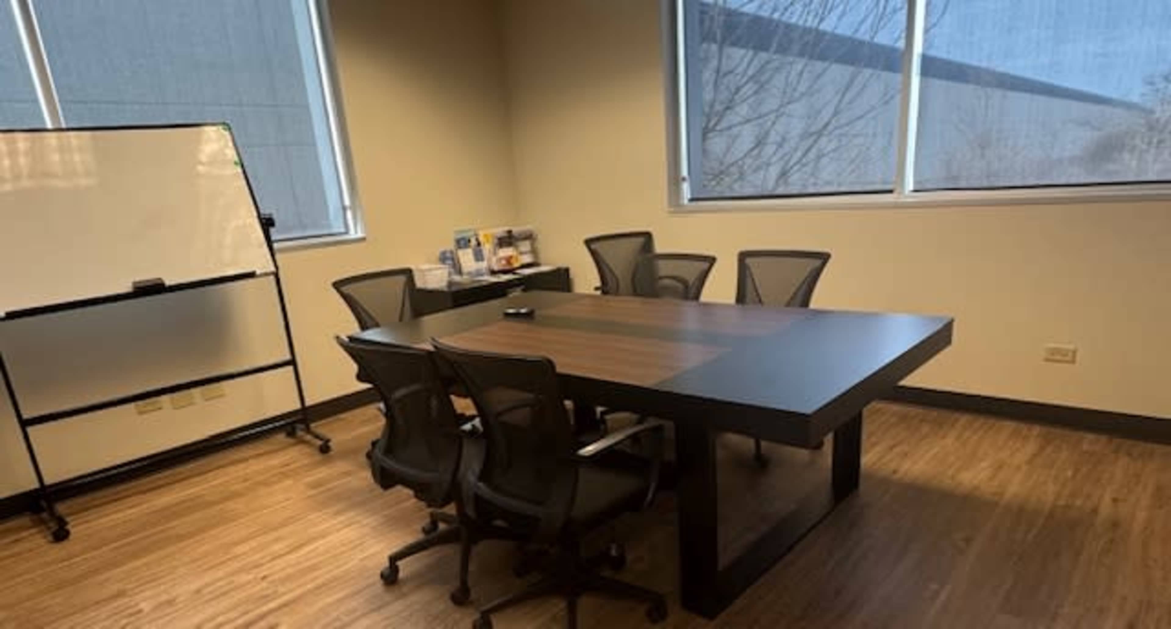 The image shows a modern conference room with a large table, four black chairs, a whiteboard, and shelves containing materials along the wall.
