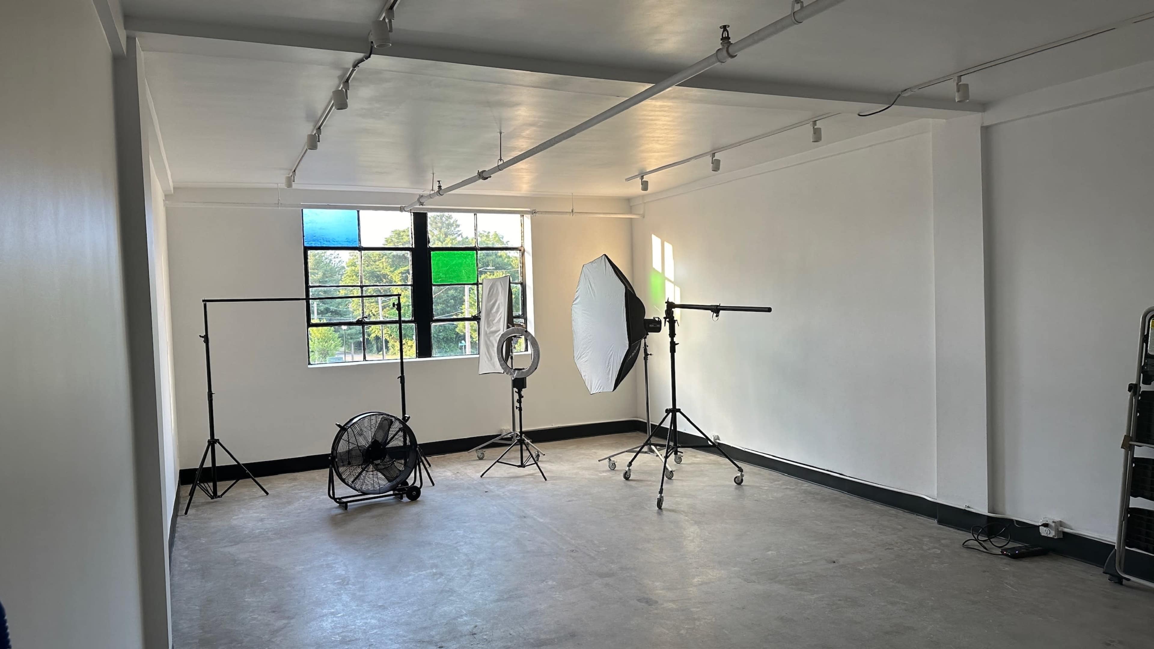A spacious, empty room with large windows featuring colored panes, a fan, and photography equipment set up for a shoot.