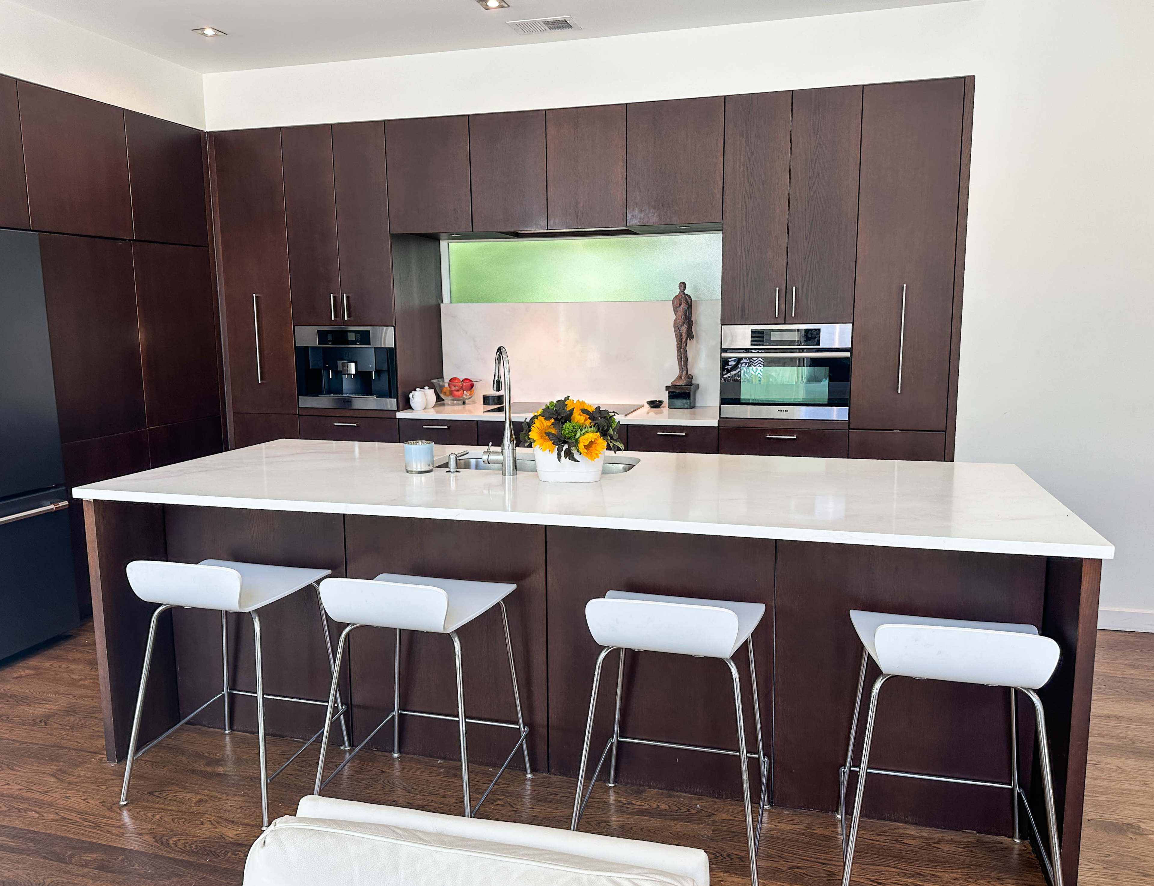 The image shows a modern kitchen with dark wood cabinetry, a large white island countertop, and four white stools.