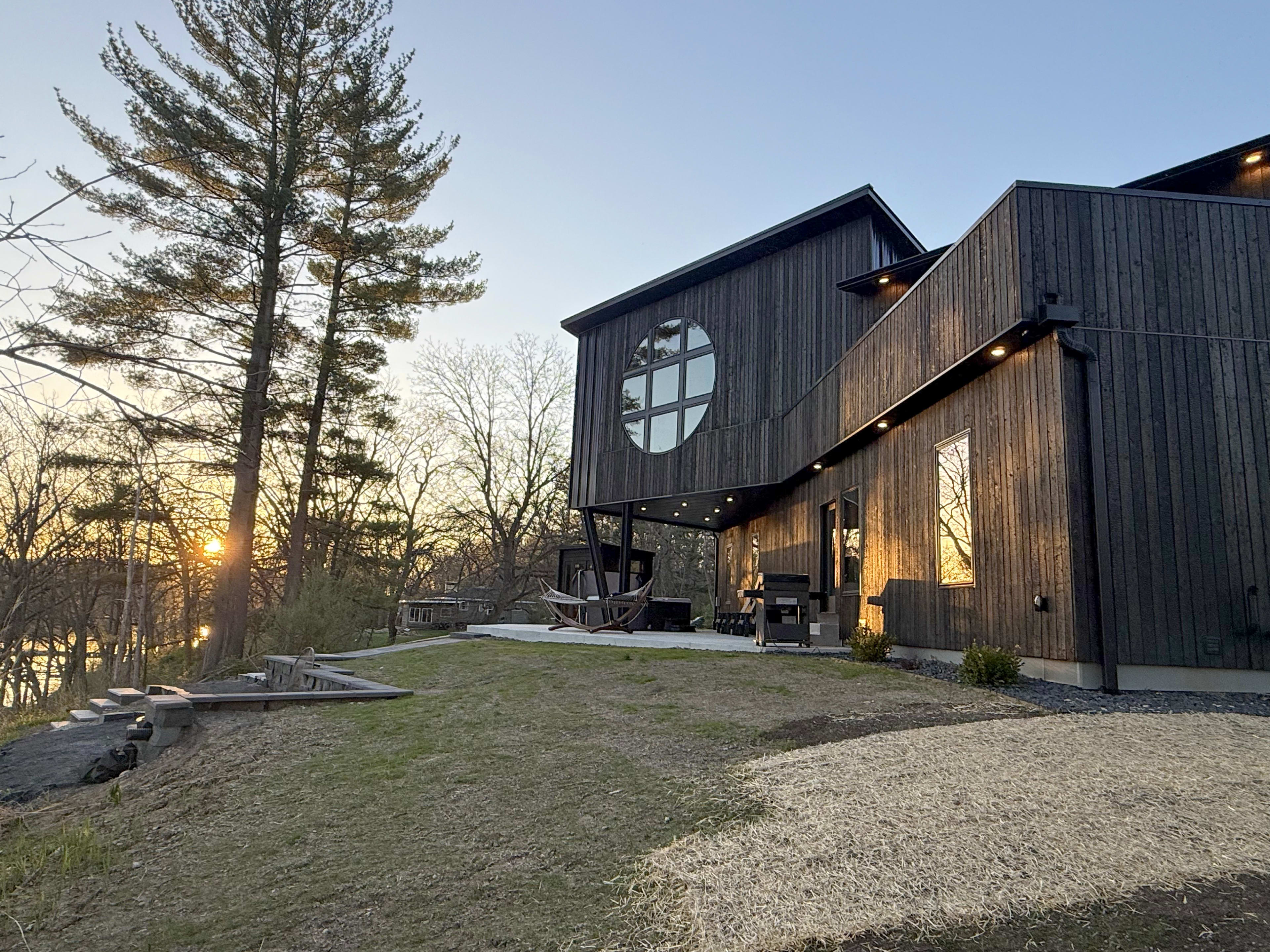 A modern wooden house with large windows overlooks a sunset near a body of water, surrounded by trees and a grassy yard.