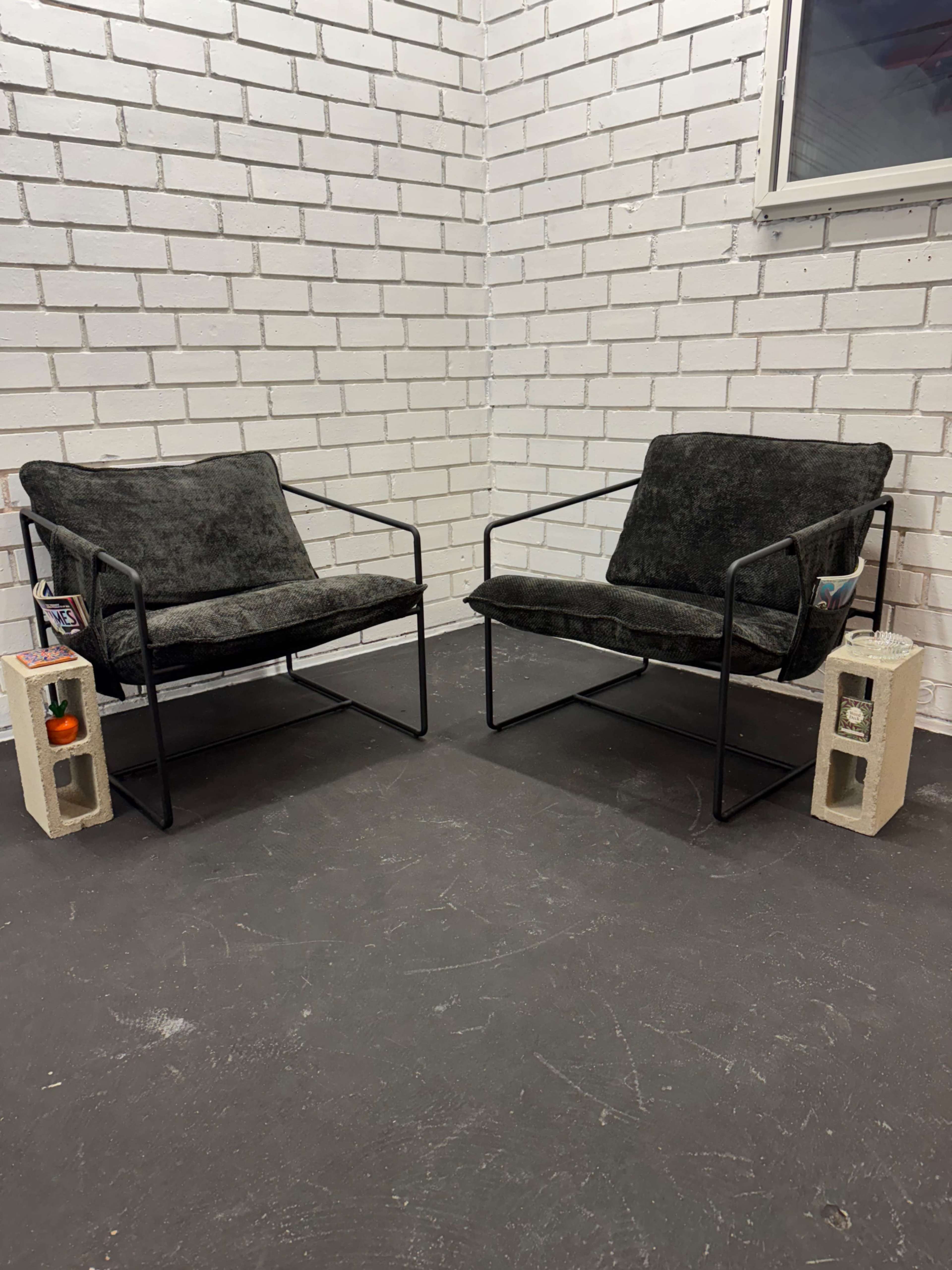 Two modern chairs with black fabric upholstery and metal frames are placed opposite each other in a room with white brick walls.