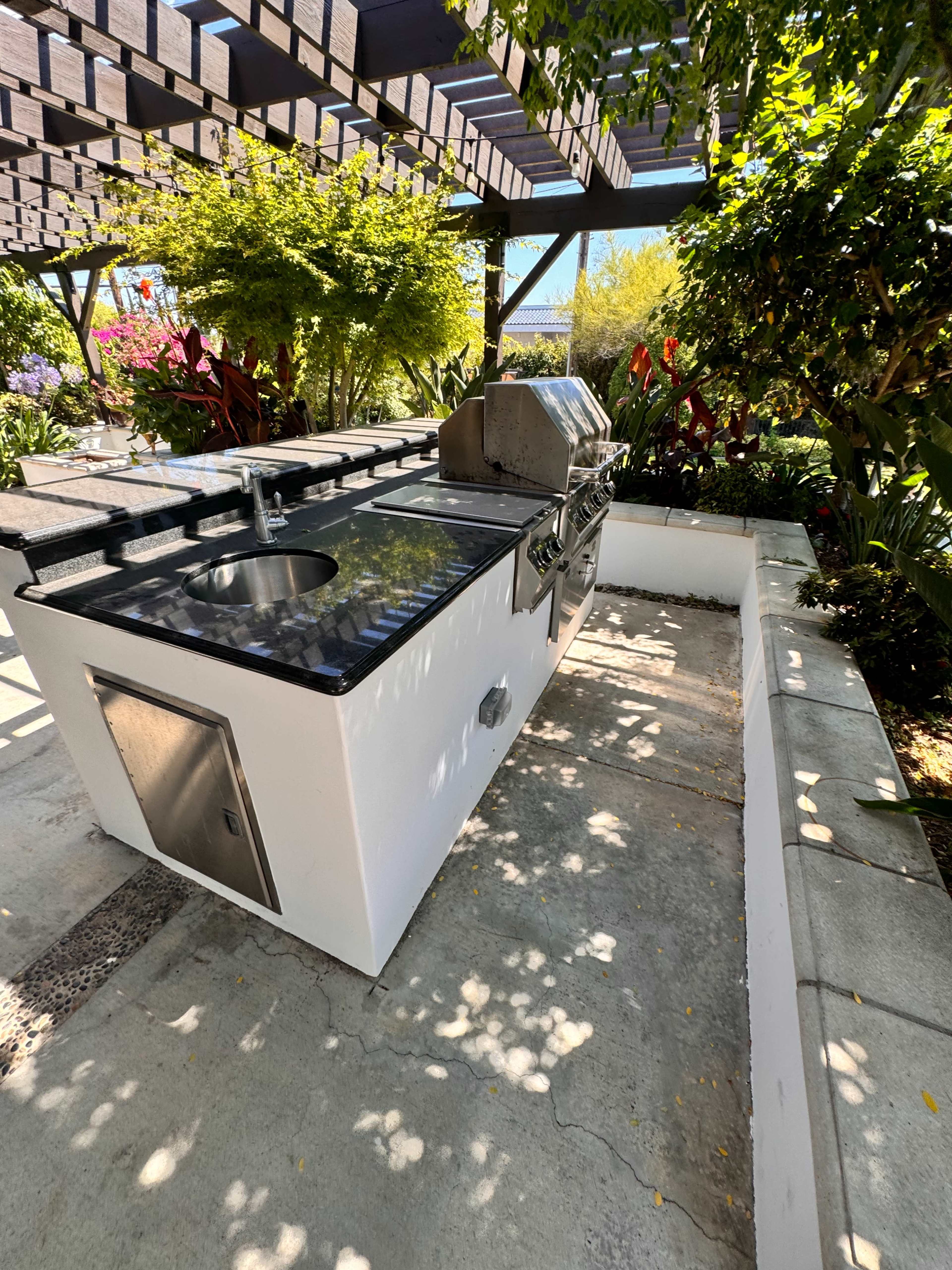 The image shows an outdoor kitchen with a built-in grill, sink, and stone countertop surrounded by lush greenery and a pergola overhead.
