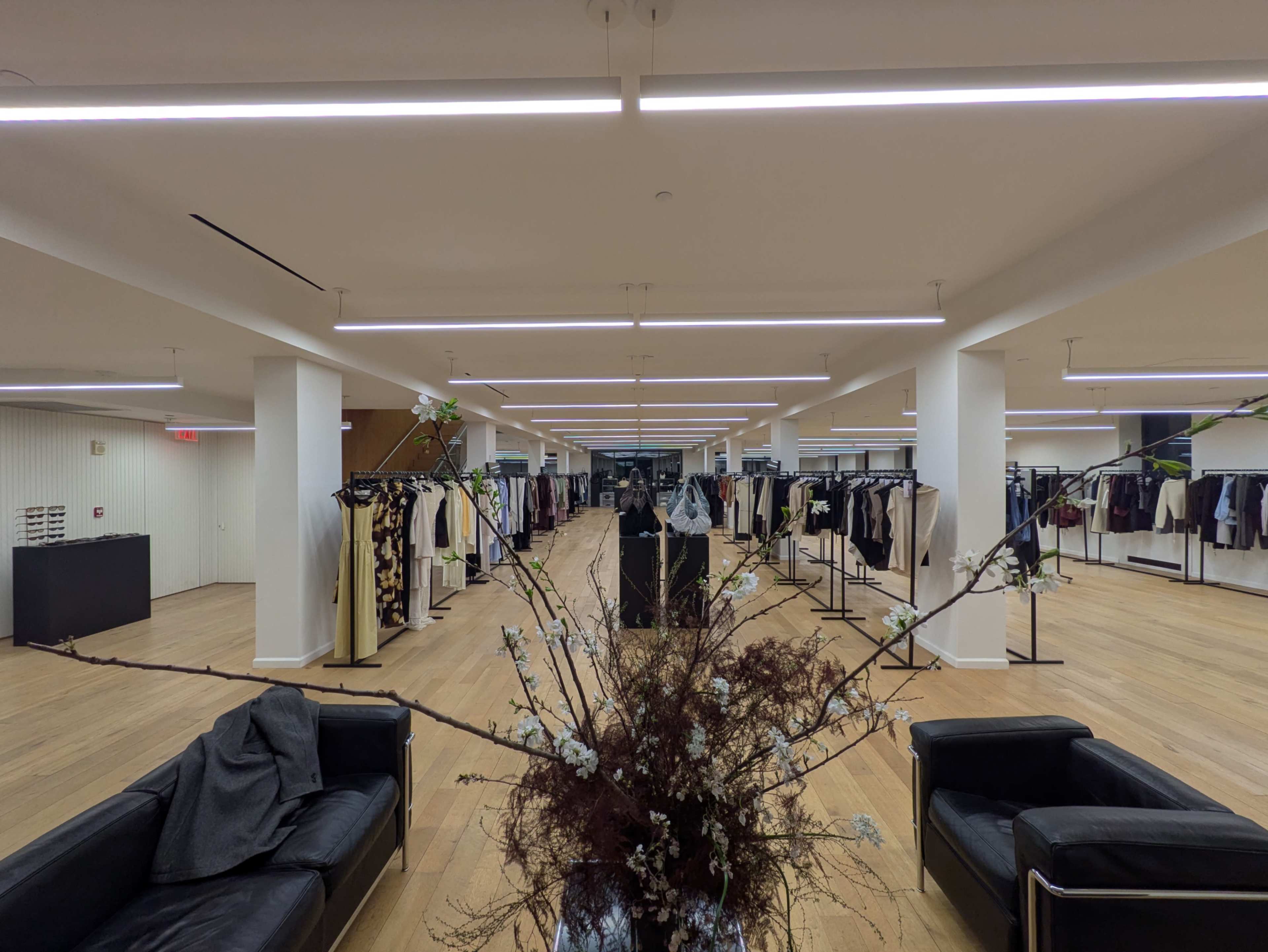 The image shows a spacious clothing store with clothing racks displaying various garments, surrounded by simple furniture and decorative plants.