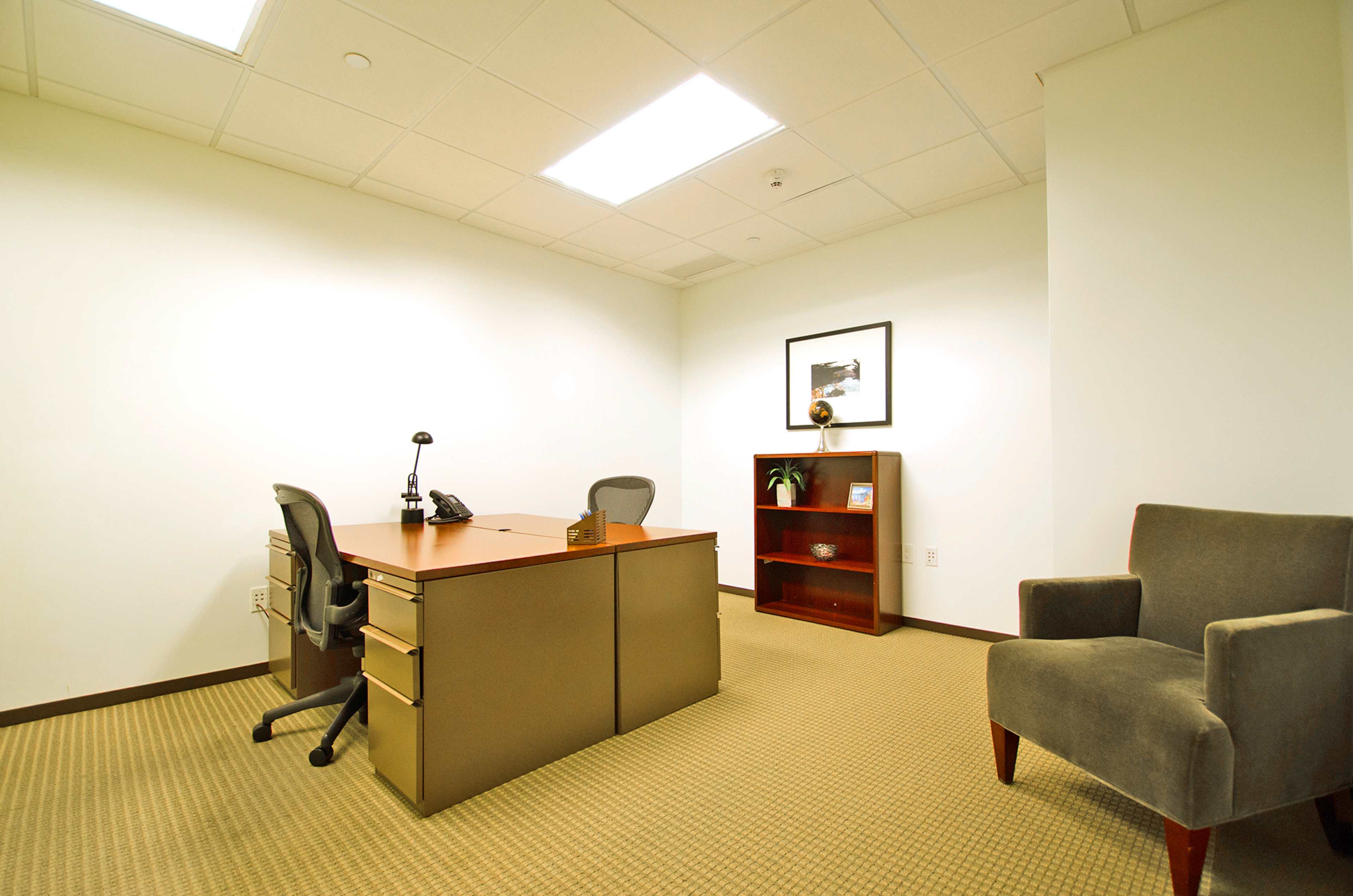 A modern office space featuring a desk with a chair, a telephone, a small bookshelf, and a single armchair against a plain wall.