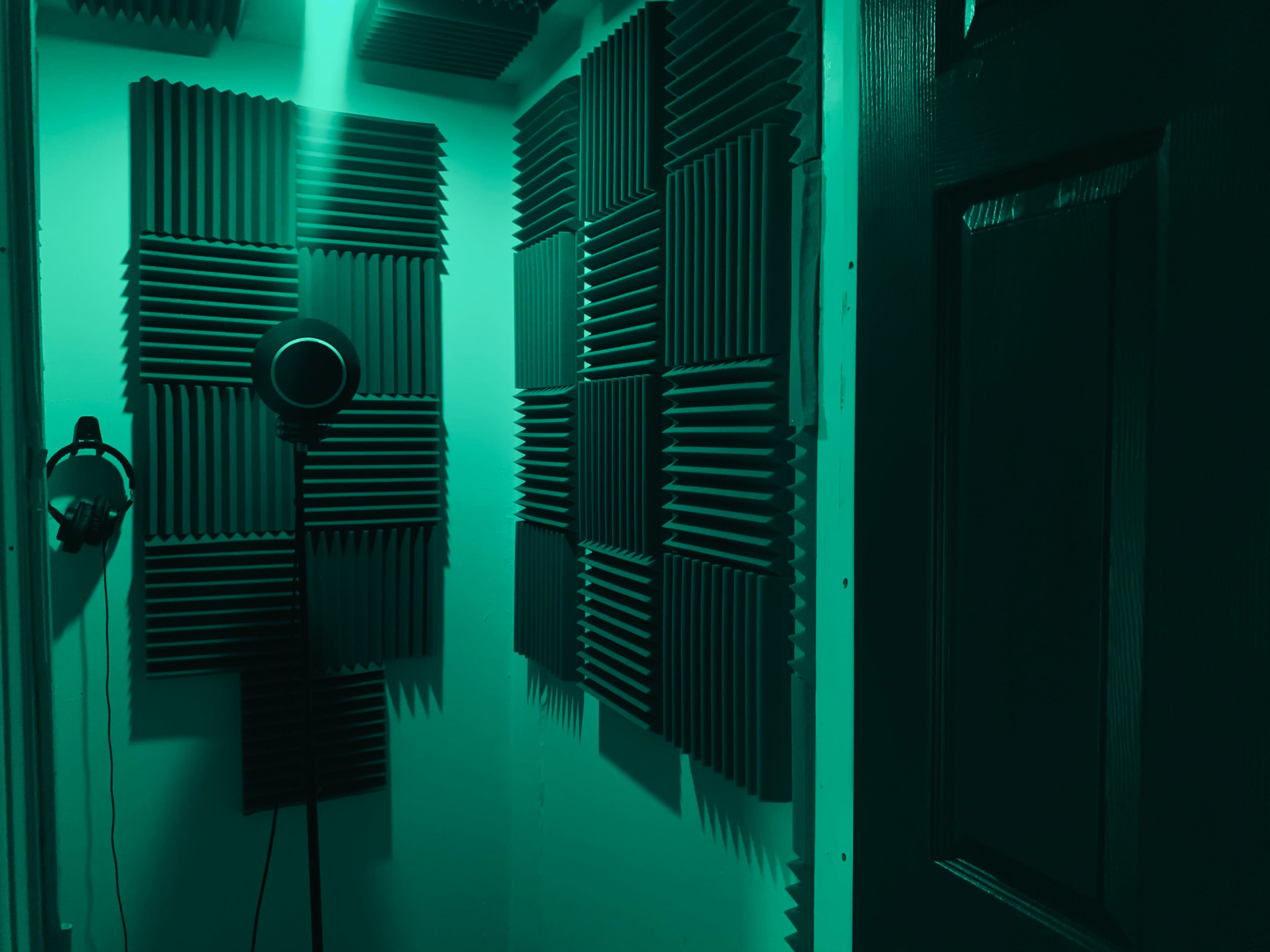 A small recording booth with soundproof foam panels on the walls, a microphone on a stand, and headphones hanging on the wall.