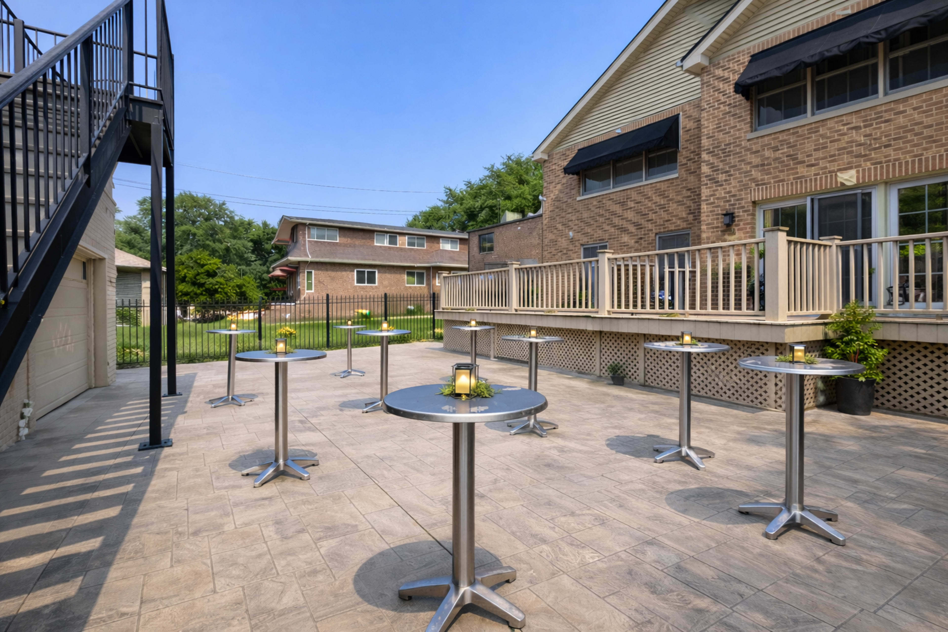 The image shows a patio area with several round tables arranged neatly, surrounded by a fence and a two-story brick building.