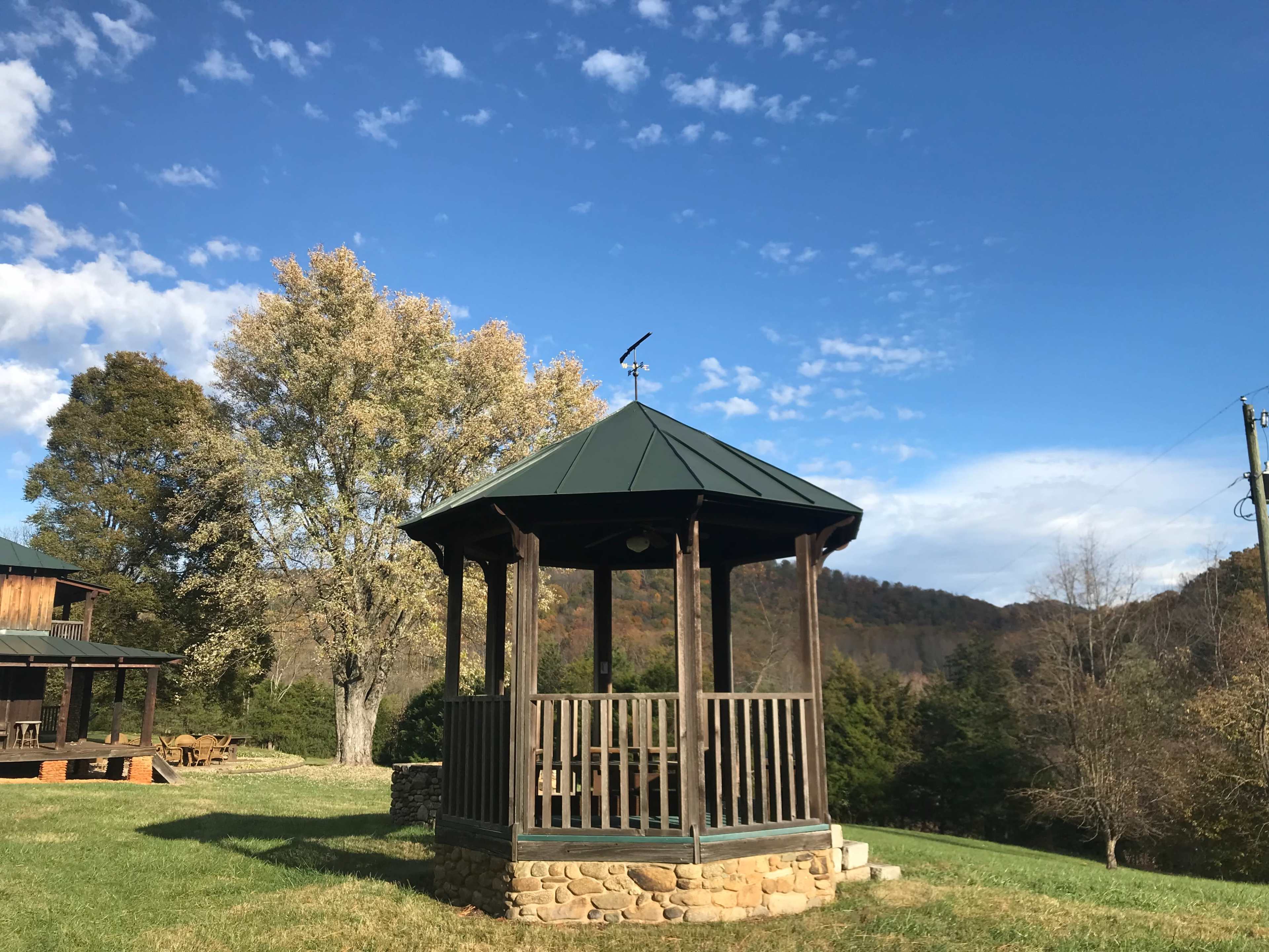 A wooden gazebo with a green roof is situated in a grassy area, surrounded by trees and under a clear blue sky.