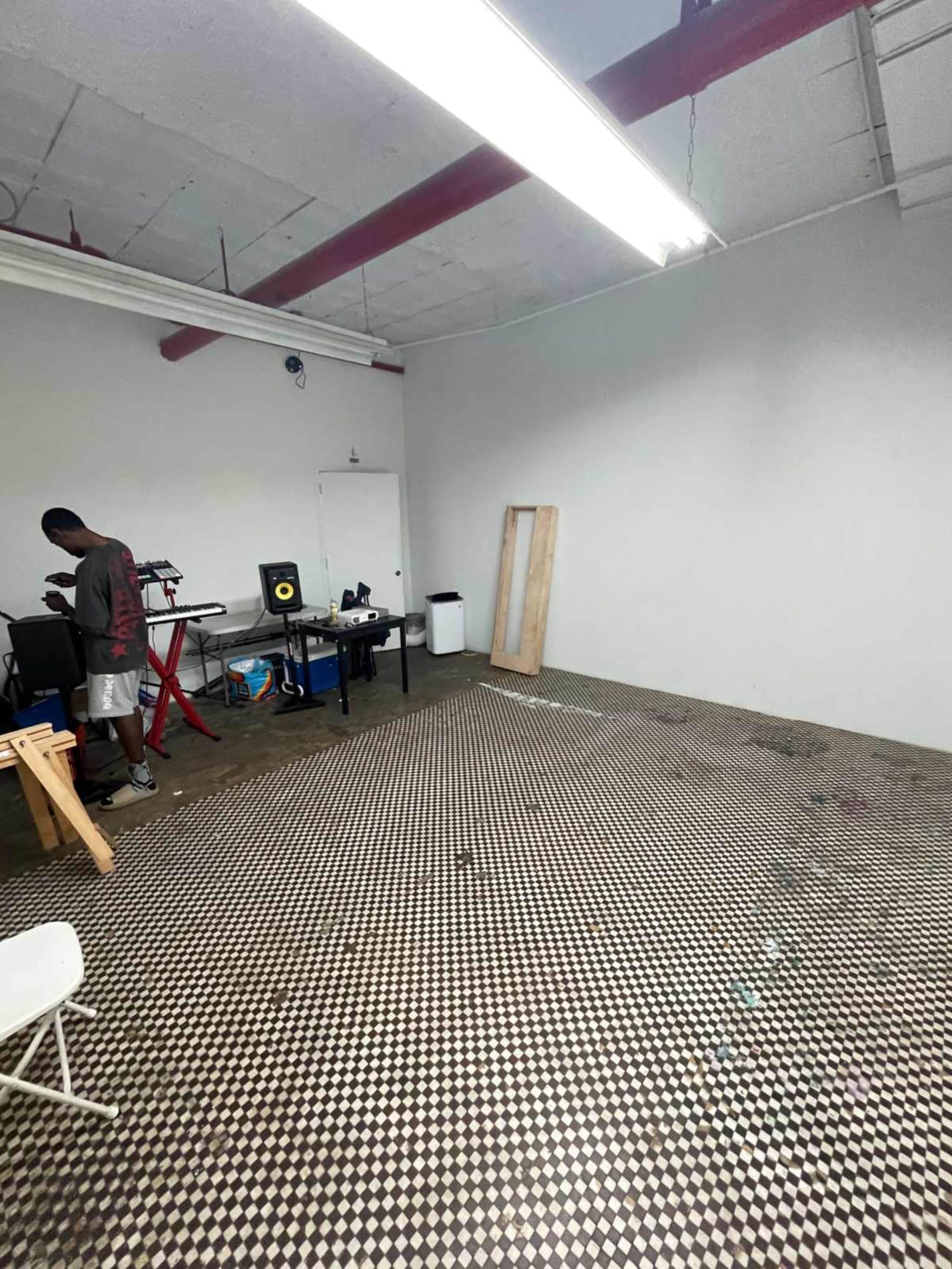 A person stands in a spacious room with a checkered floor, equipped with musical instruments and furniture along the walls.