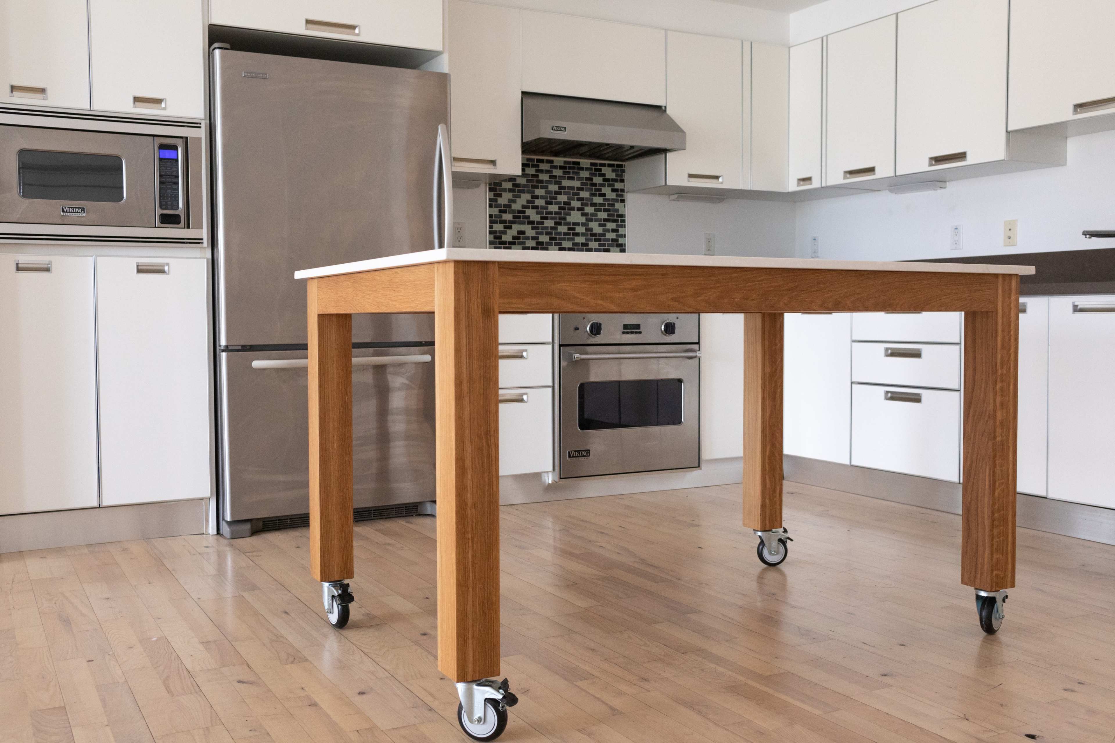 A wooden kitchen table on wheels positioned in a modern kitchen with stainless steel appliances and white cabinets.