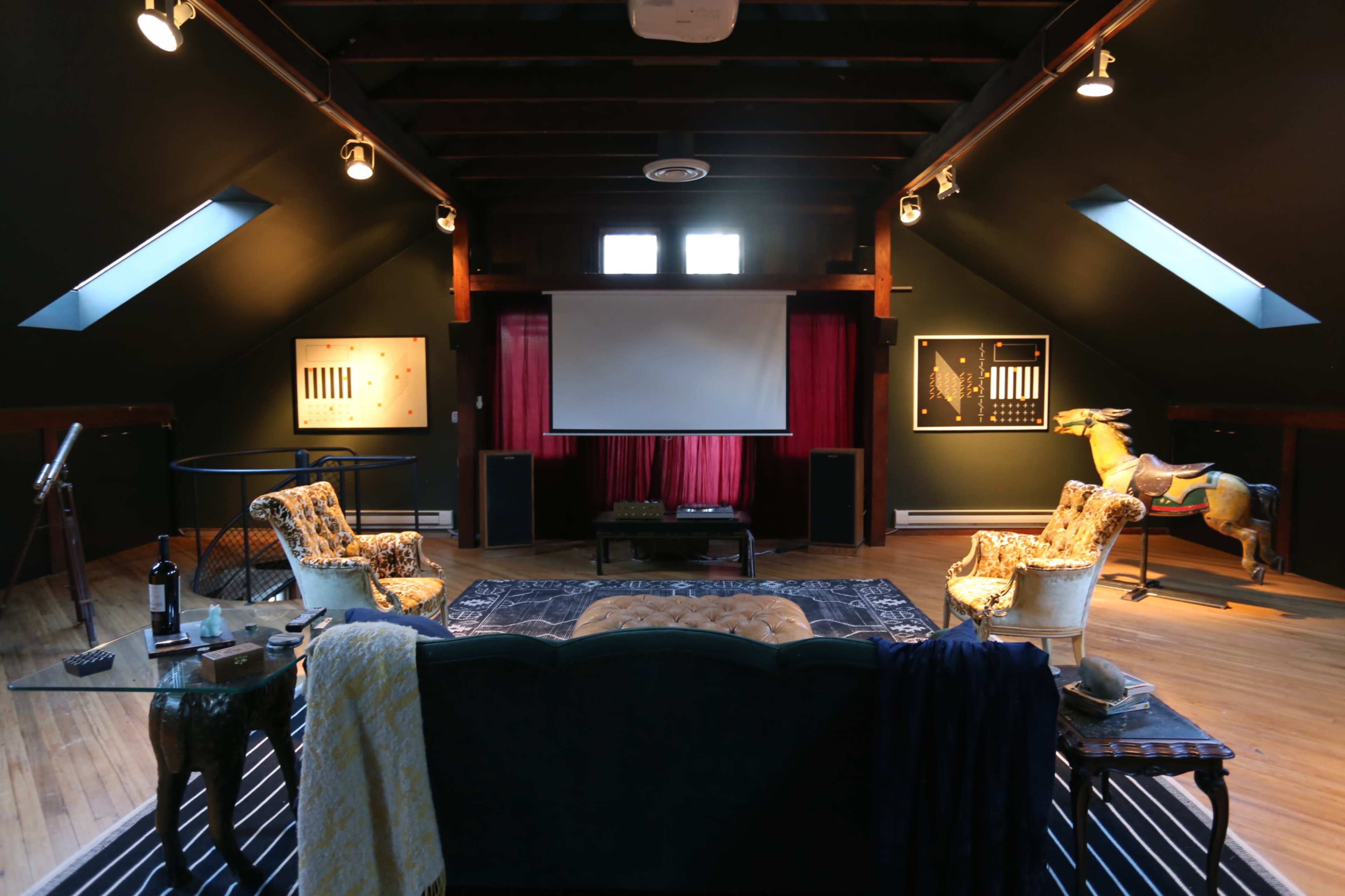 A spacious attic room with a large screen, two upholstered chairs, a coffee table, and artworks on the walls.