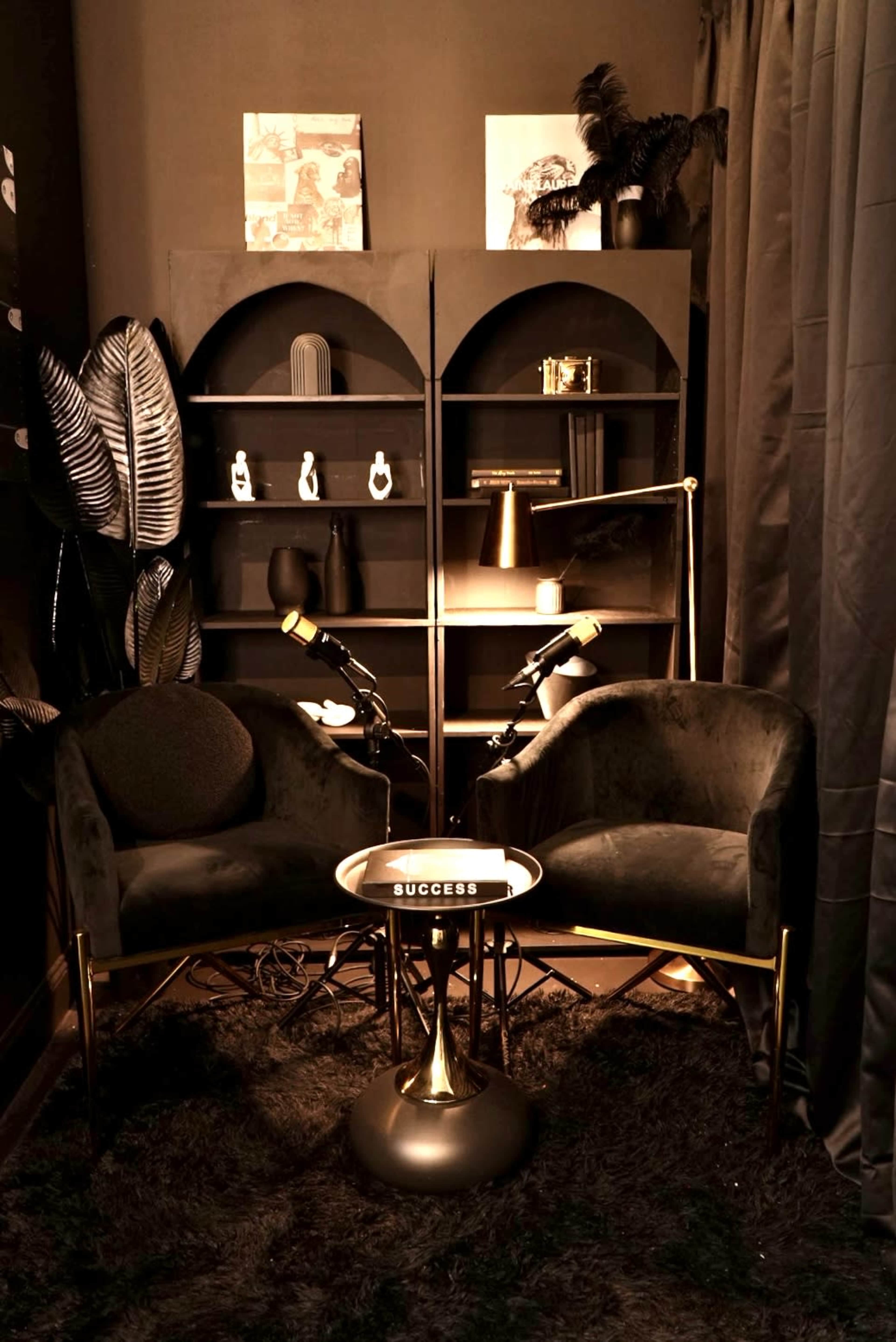 The image shows a cozy corner with two plush chairs, a small circular table labeled "SUCCESS," and a stylish bookshelf, all set against dark walls and draped curtains.