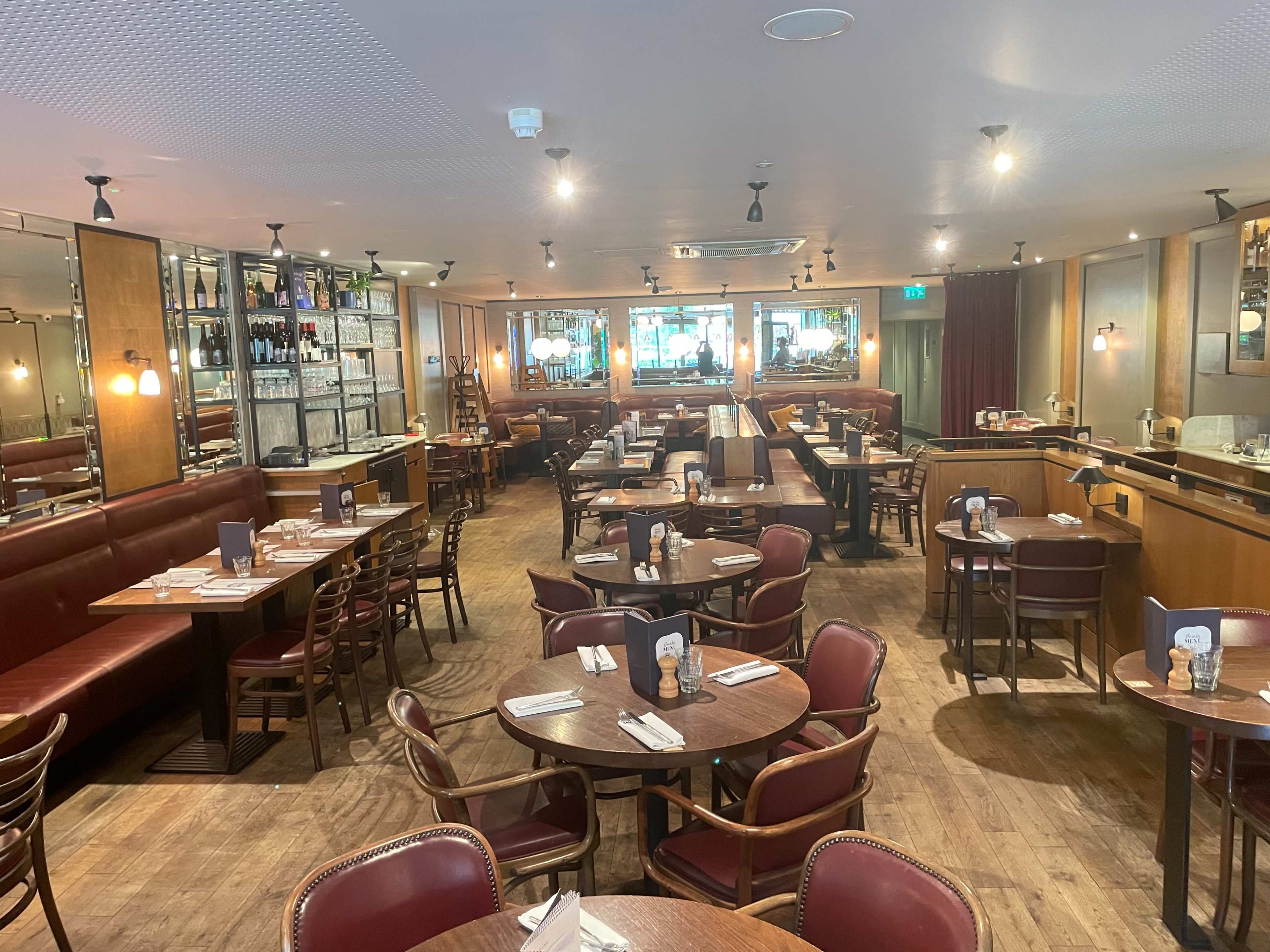 The image shows an empty restaurant featuring wooden tables and chairs, red leather booths, and a bar area in the background.