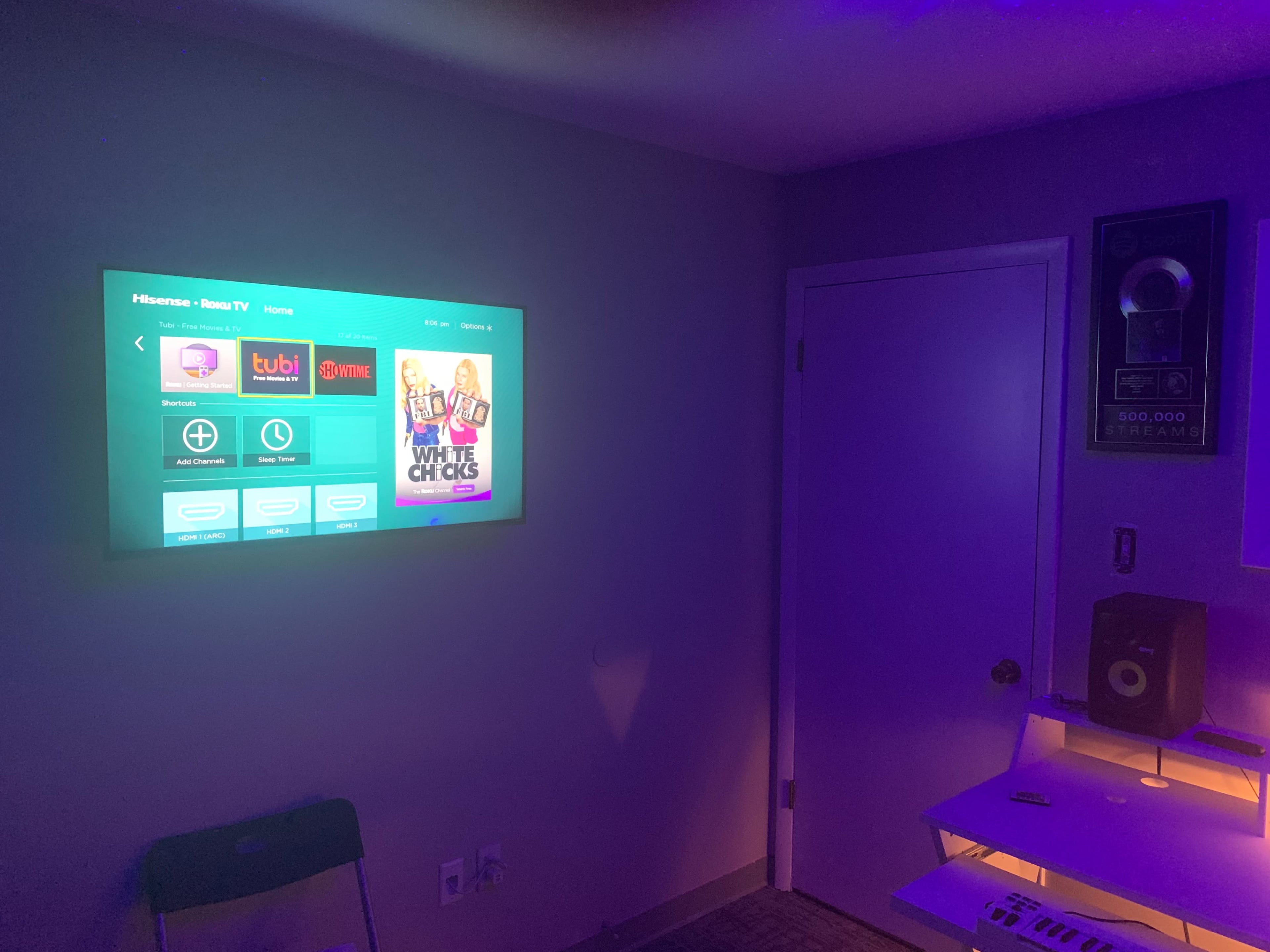 A wall-mounted TV displaying streaming app options in a dimly lit room with a door and a speaker on a desk.