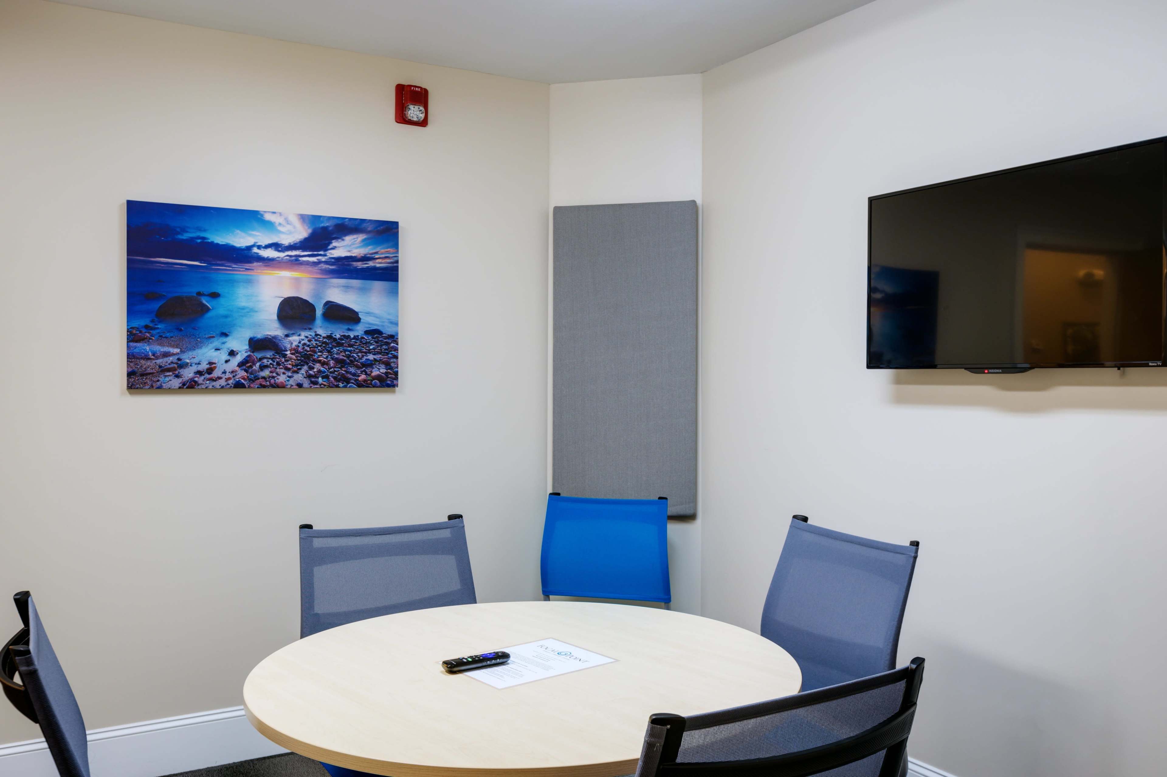 The image shows a small meeting room with a round table, four chairs, a wall-mounted television, and a wall art piece depicting a beach scene.