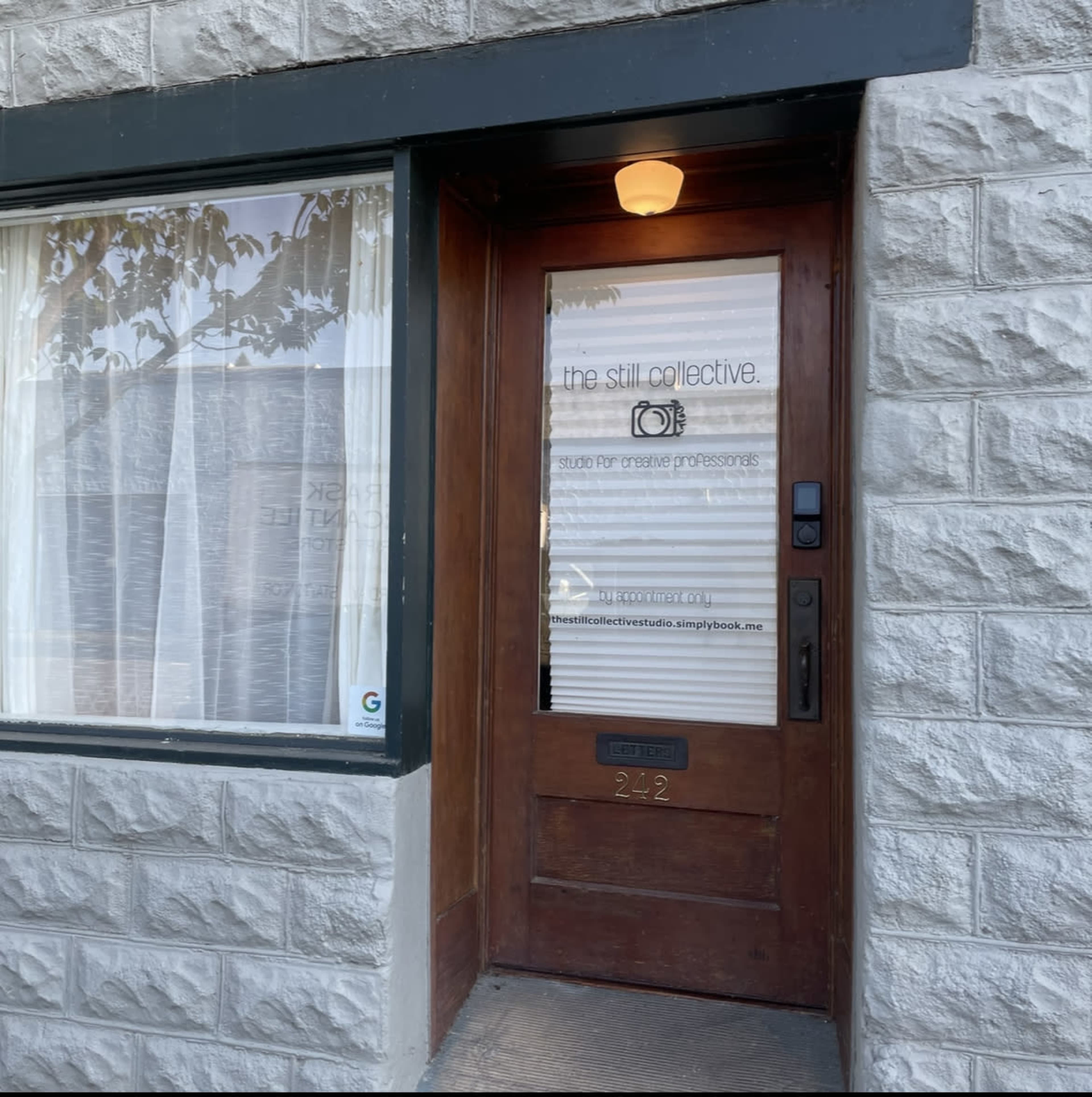 A wooden door with an etched sign reading "the still collective" and an indicator for "studio for creative professionals," surrounded by stone walls and a large window with sheer curtains.