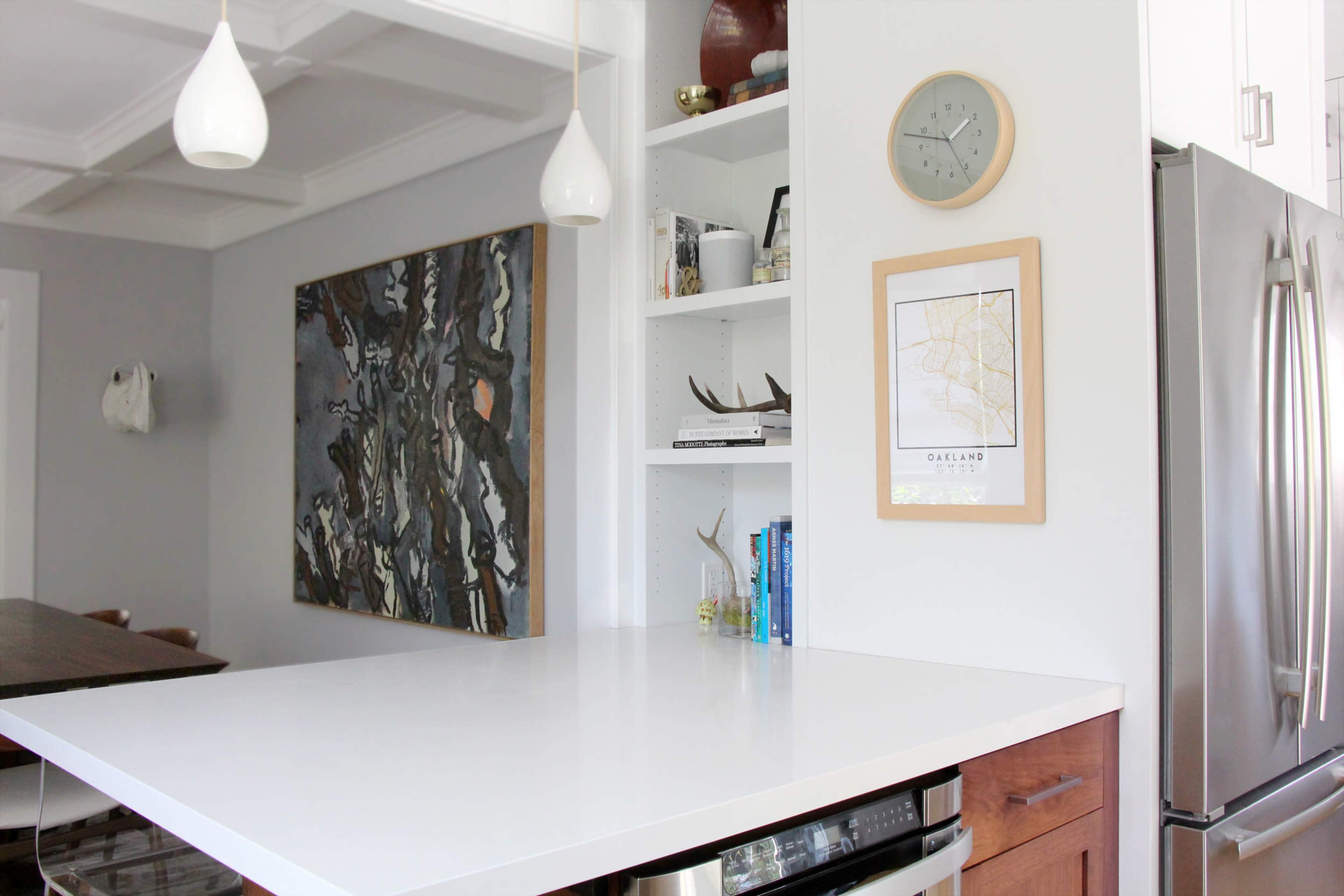 A modern kitchen features a white countertop, shelves with books and decor, and a wall art piece beside a clock.