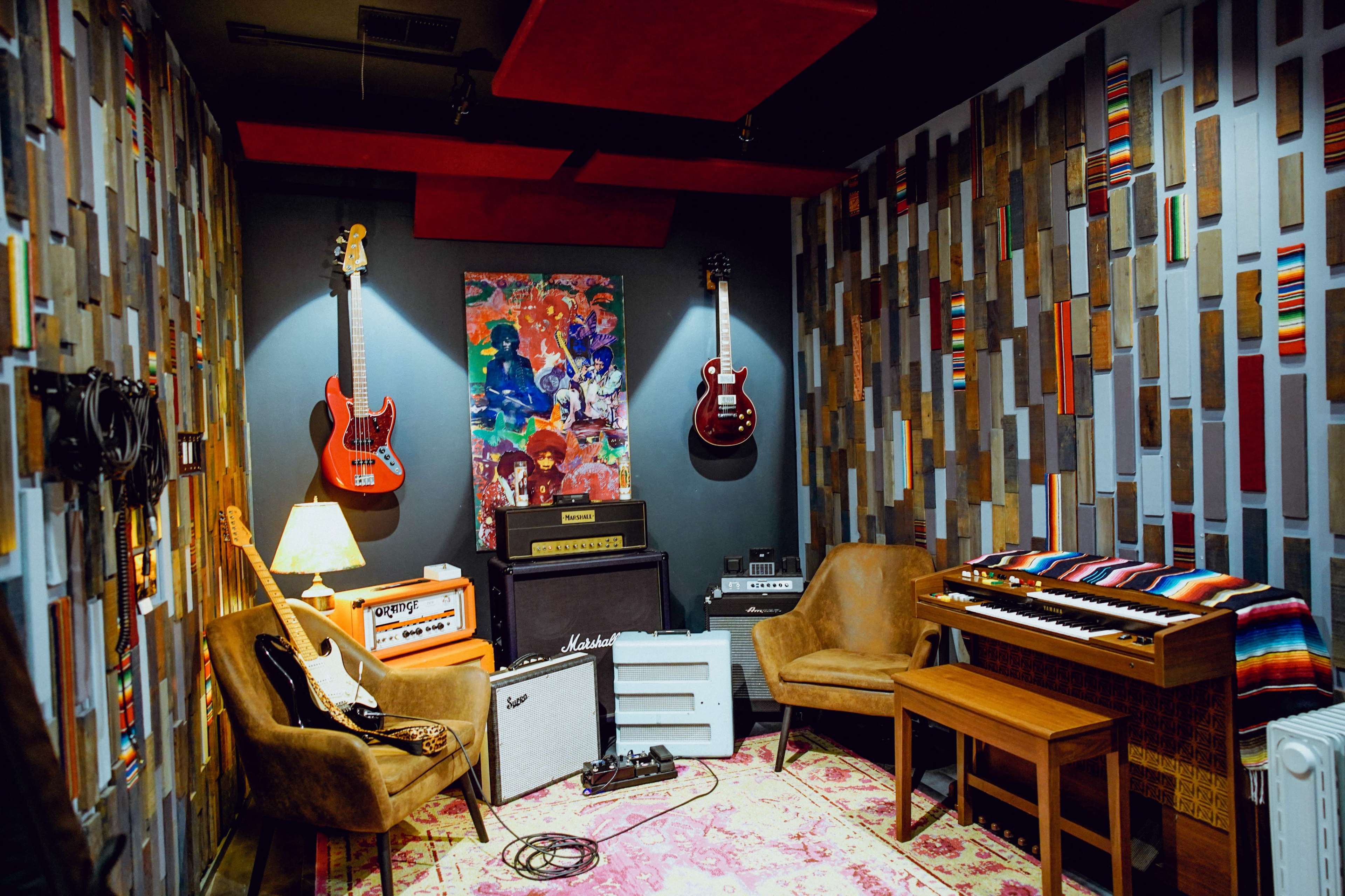The image shows a music studio corner featuring two electric guitars on the wall, a vintage keyboard, amplifiers, and comfortable chairs, all set against a colorful patterned wall.