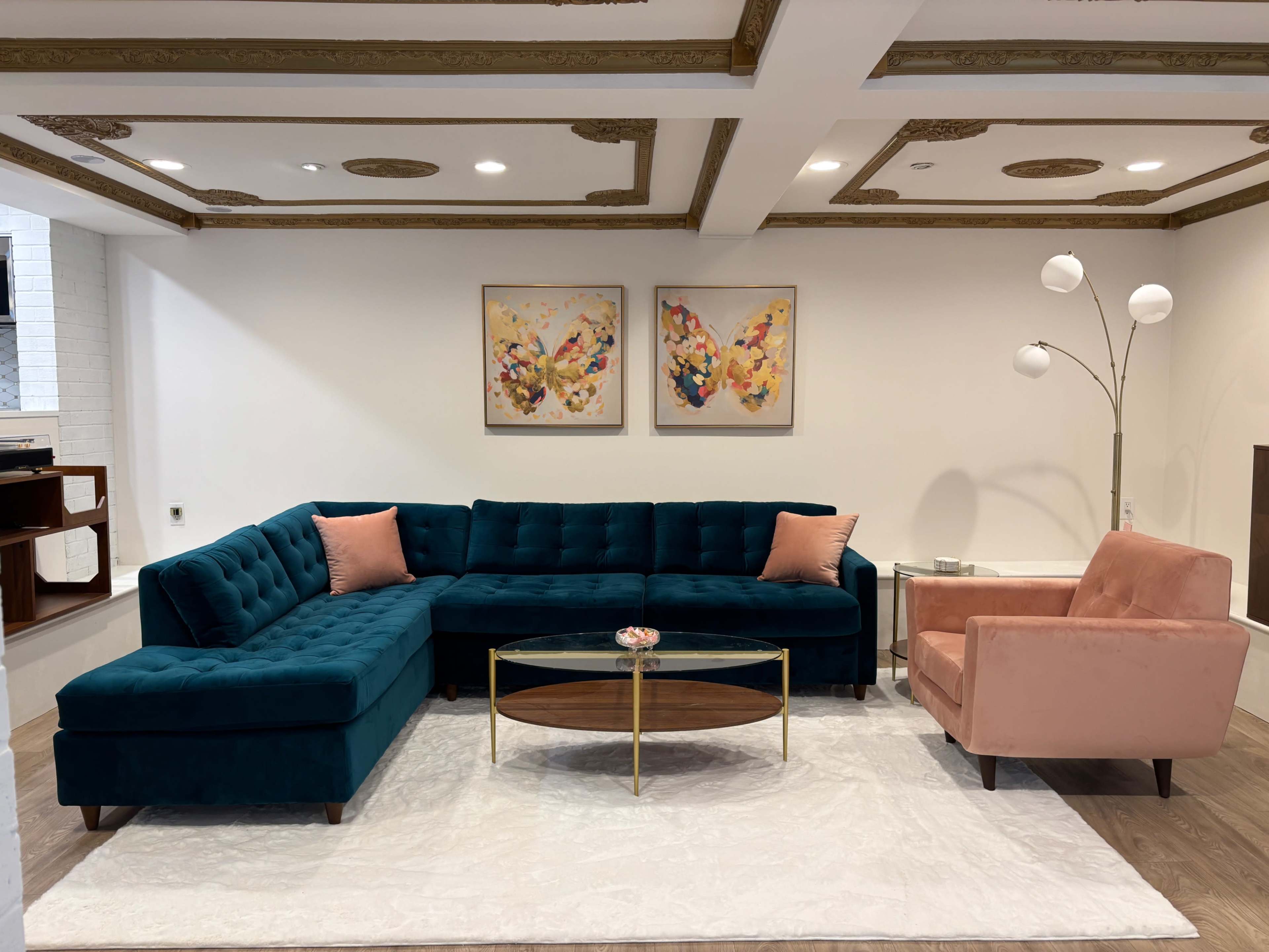 The image shows a modern living room featuring a teal sectional sofa with pink accents, a glass coffee table, and a light pink armchair under a decorative ceiling.