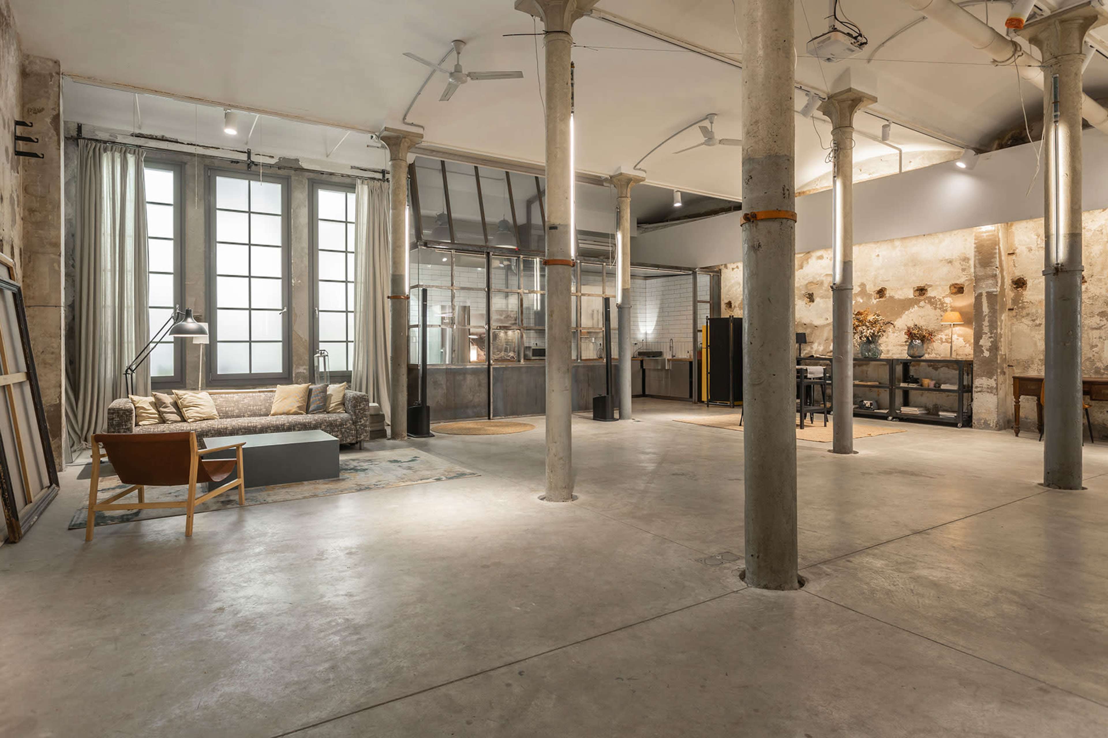 The image shows an industrial-style interior space featuring concrete pillars, large windows, and a mix of furniture including a sofa and chairs.