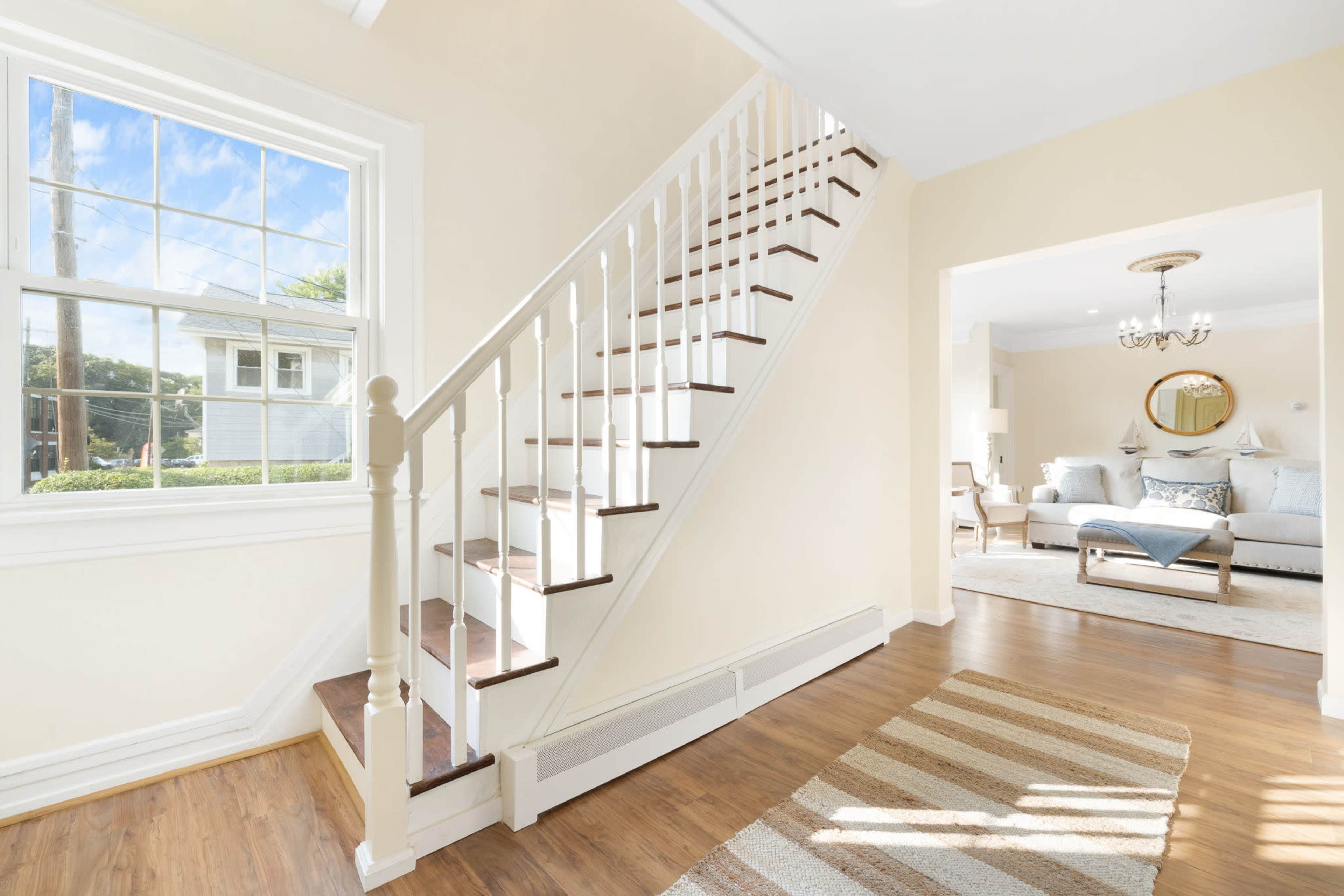 A staircase with white railings leads to an upper floor in a bright, light-colored entryway with a striped rug and a view of a living room in the background.