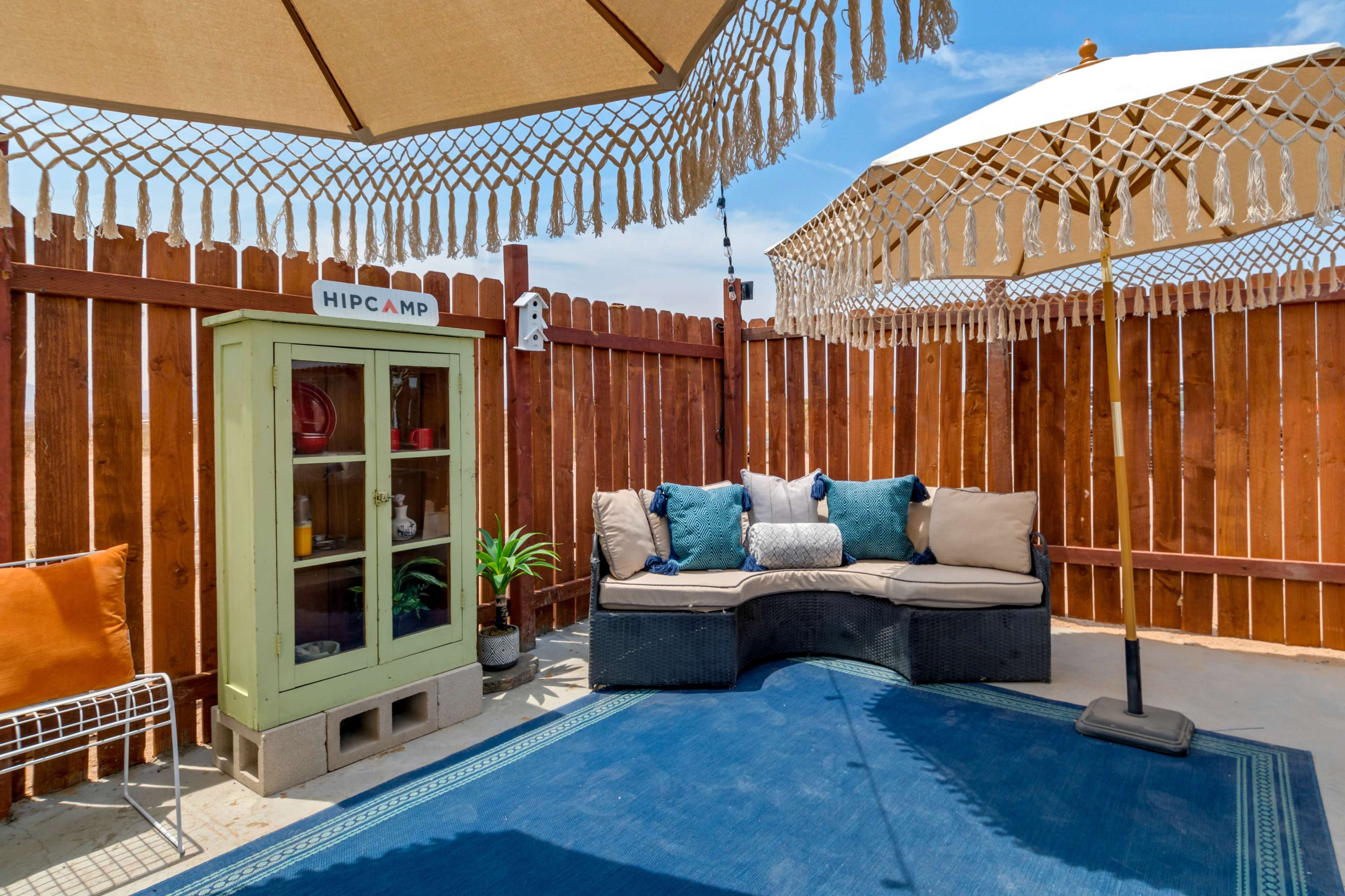 The scene features a cozy outdoor seating area with an umbrella, decorative string lights, a small cabinet labeled "HIPCAMP," and a blue rug on a sandy surface.