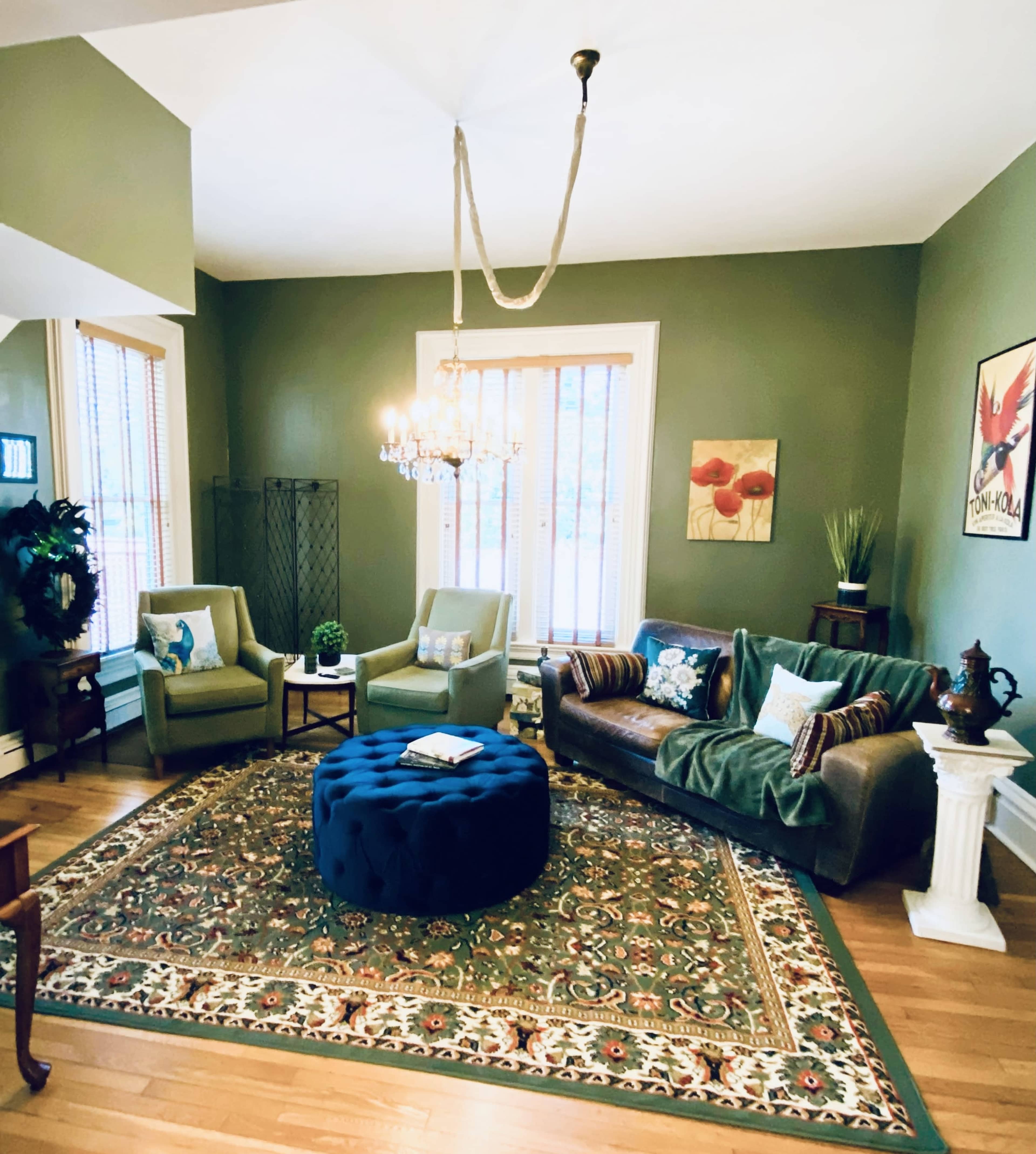 A cozy living room featuring a circular blue ottoman, a brown sofa with a throw blanket, two green chairs, and a decorative chandelier above, all set against green walls and a patterned area rug.