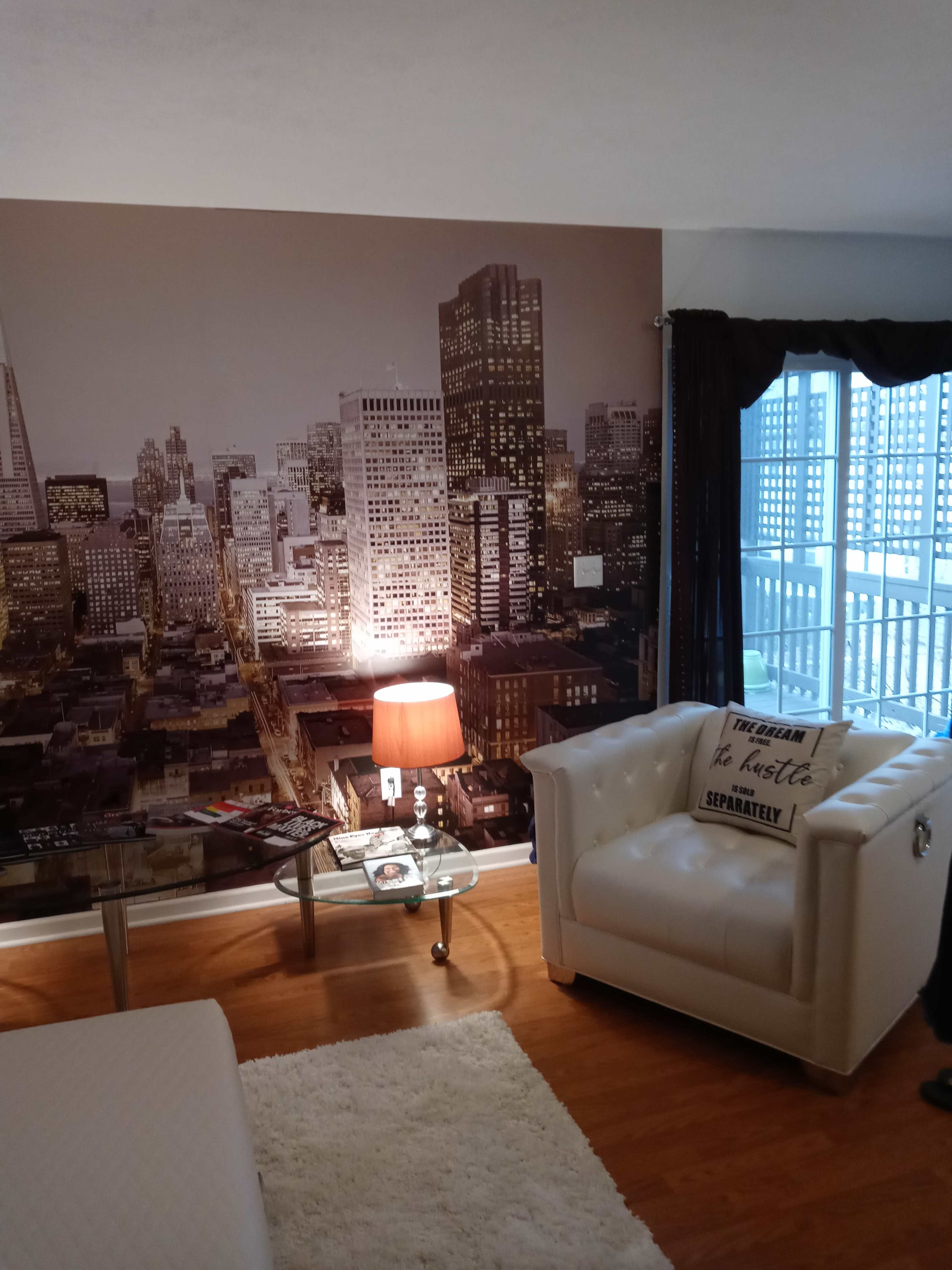The image shows a modern living space featuring a large cityscape mural, a white leather armchair, a glass coffee table with magazines, and a floor lamp.