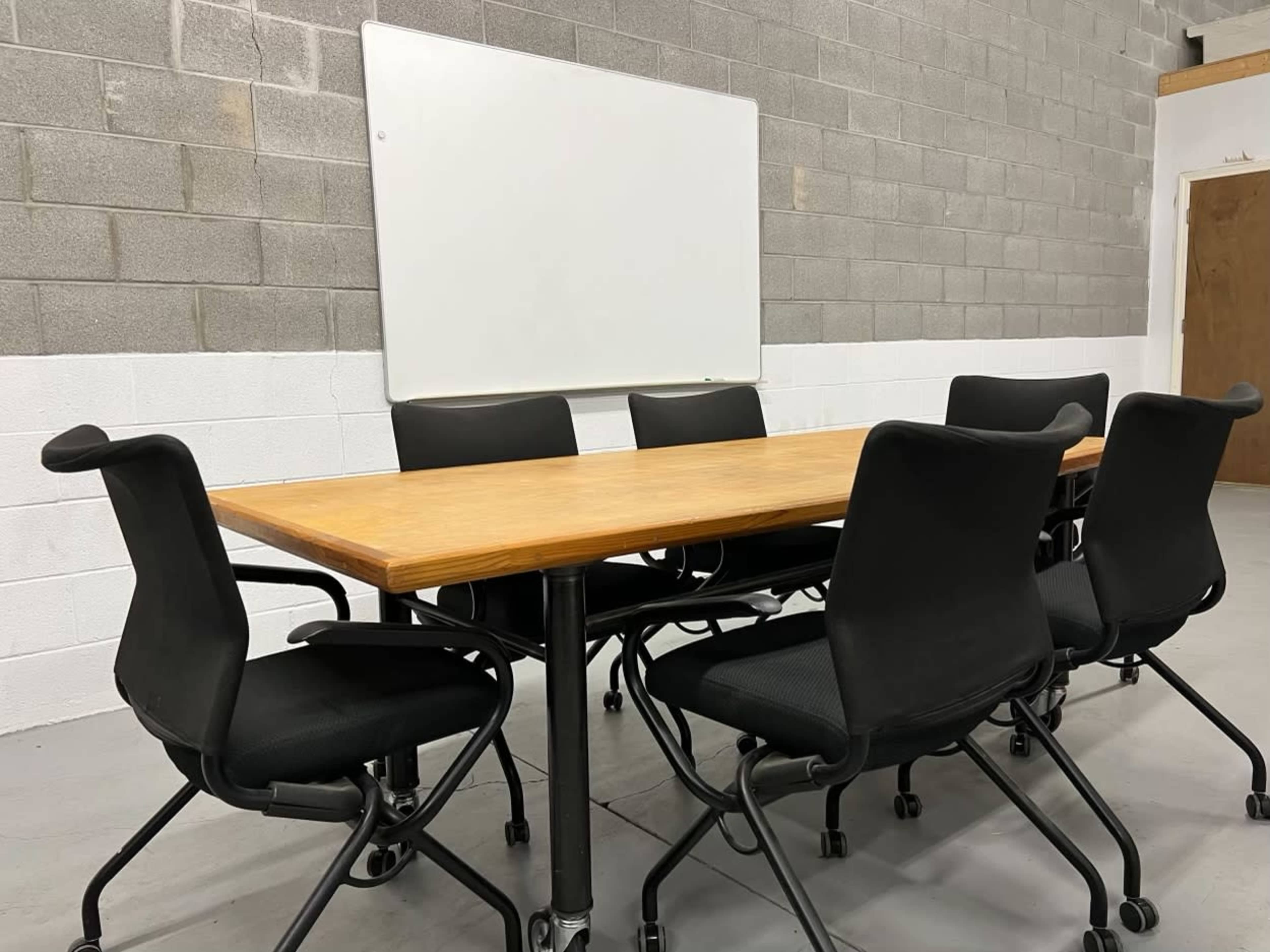 A rectangular wooden conference table is surrounded by six black rolling chairs in a room with gray block walls and a whiteboard mounted on the wall.