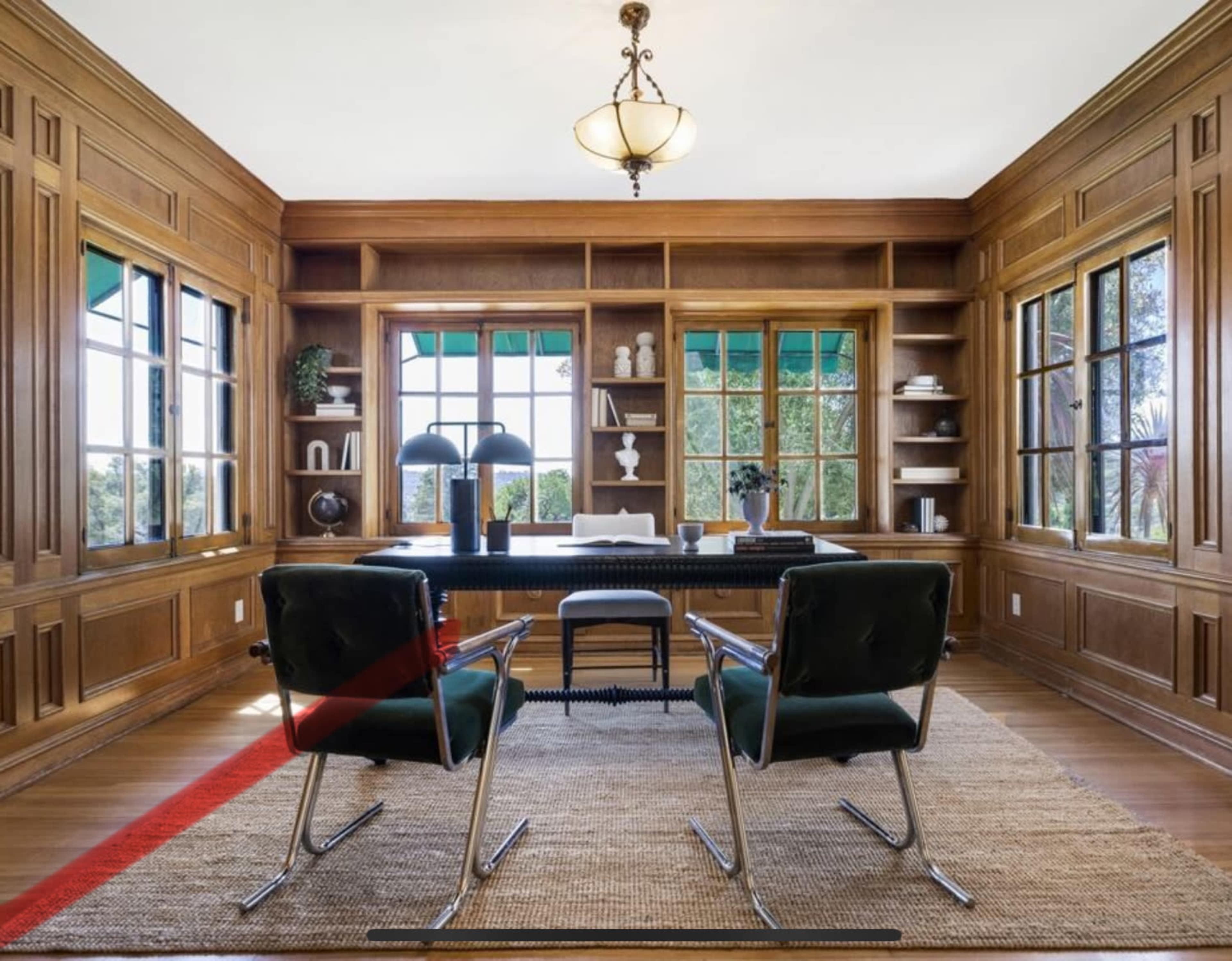 The image shows a well-appointed wooden office with two black chairs facing a desk, surrounded by large windows and built-in shelves.