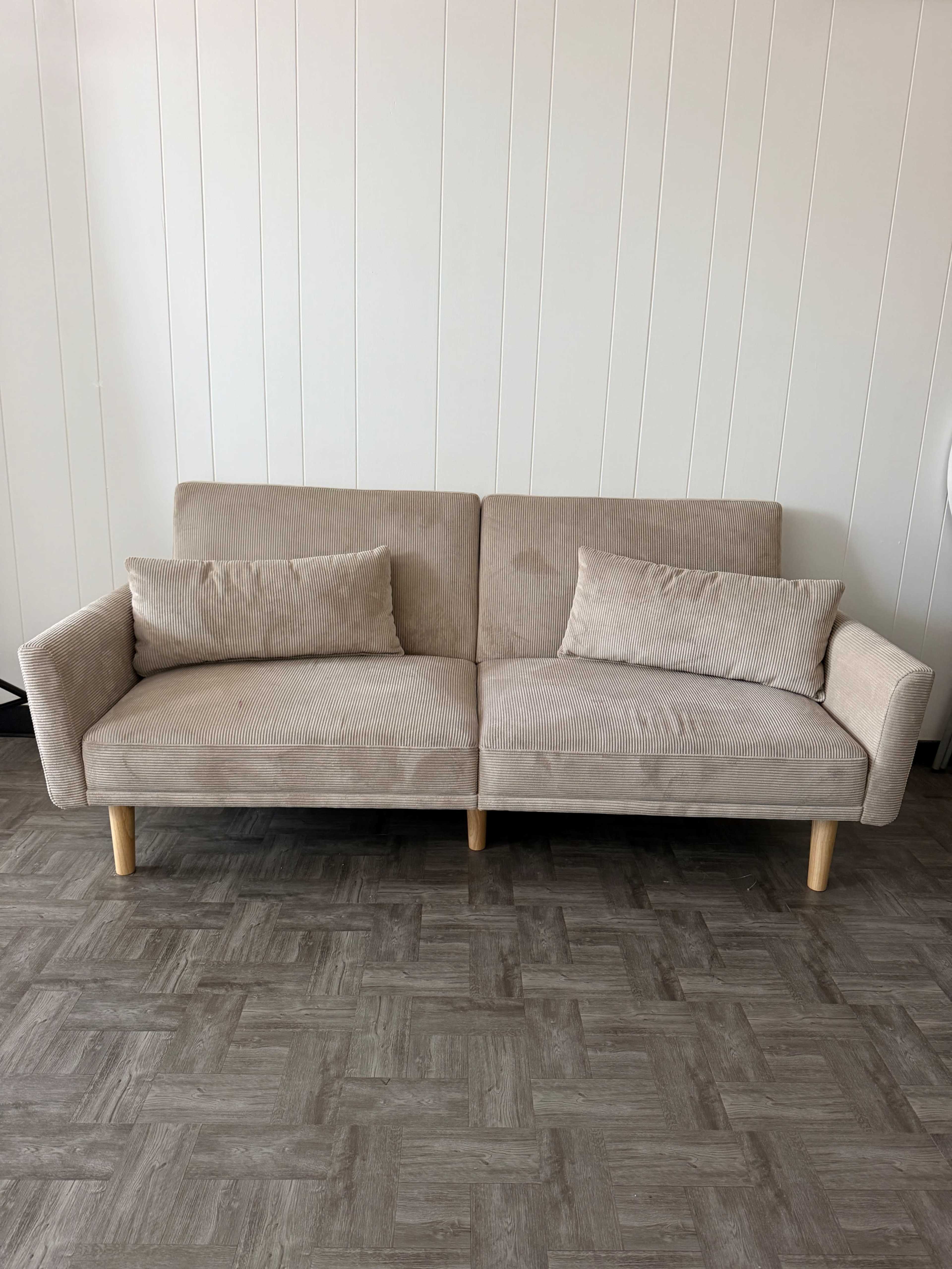 A light beige, two-seater sofa with wooden legs and two matching cushions is positioned against a white wall on a patterned floor.