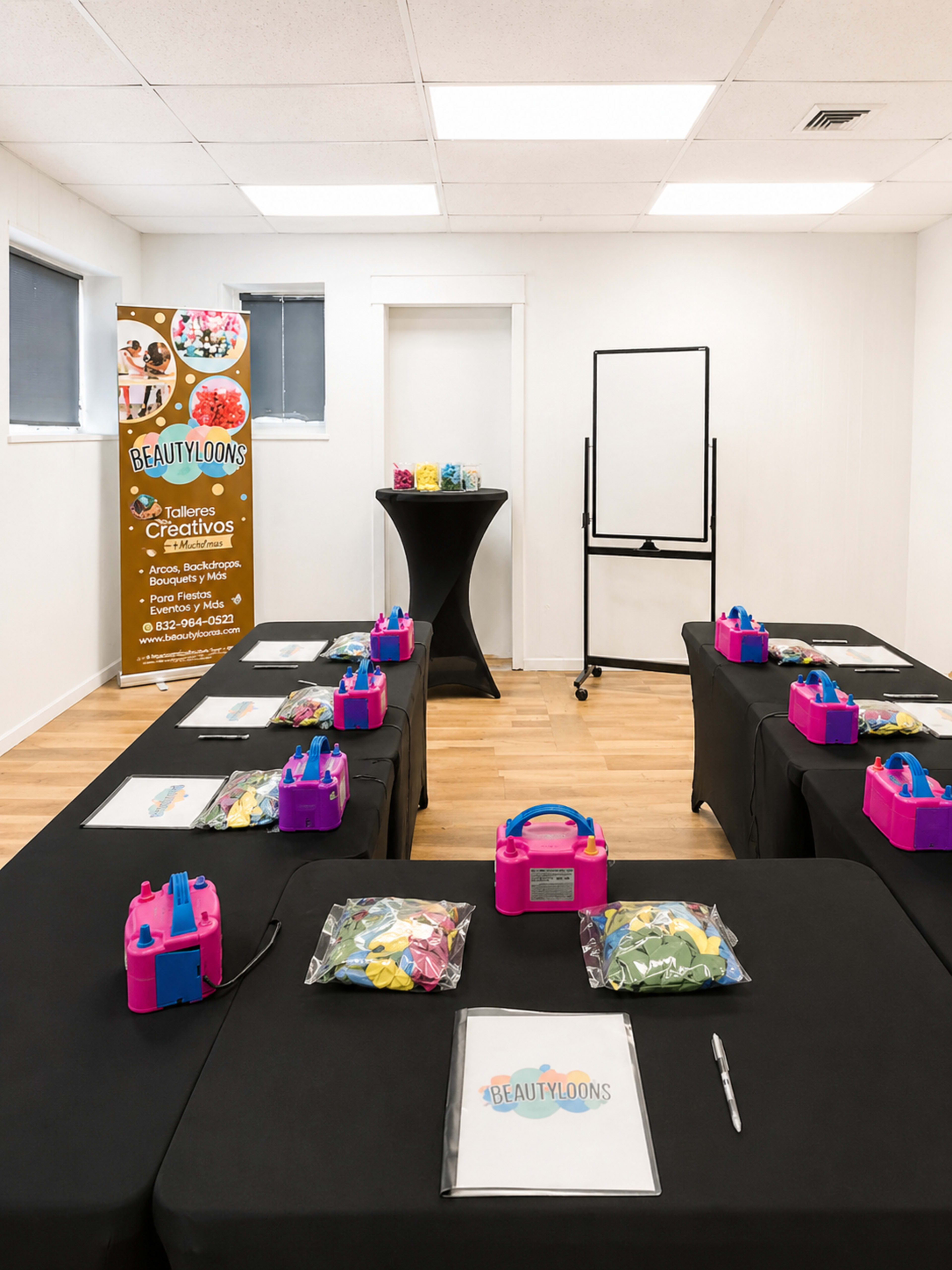 A brightly lit classroom features several tables covered with black cloths, each equipped with pink balloon kits and a sign for "BeautyLoons" in the background.