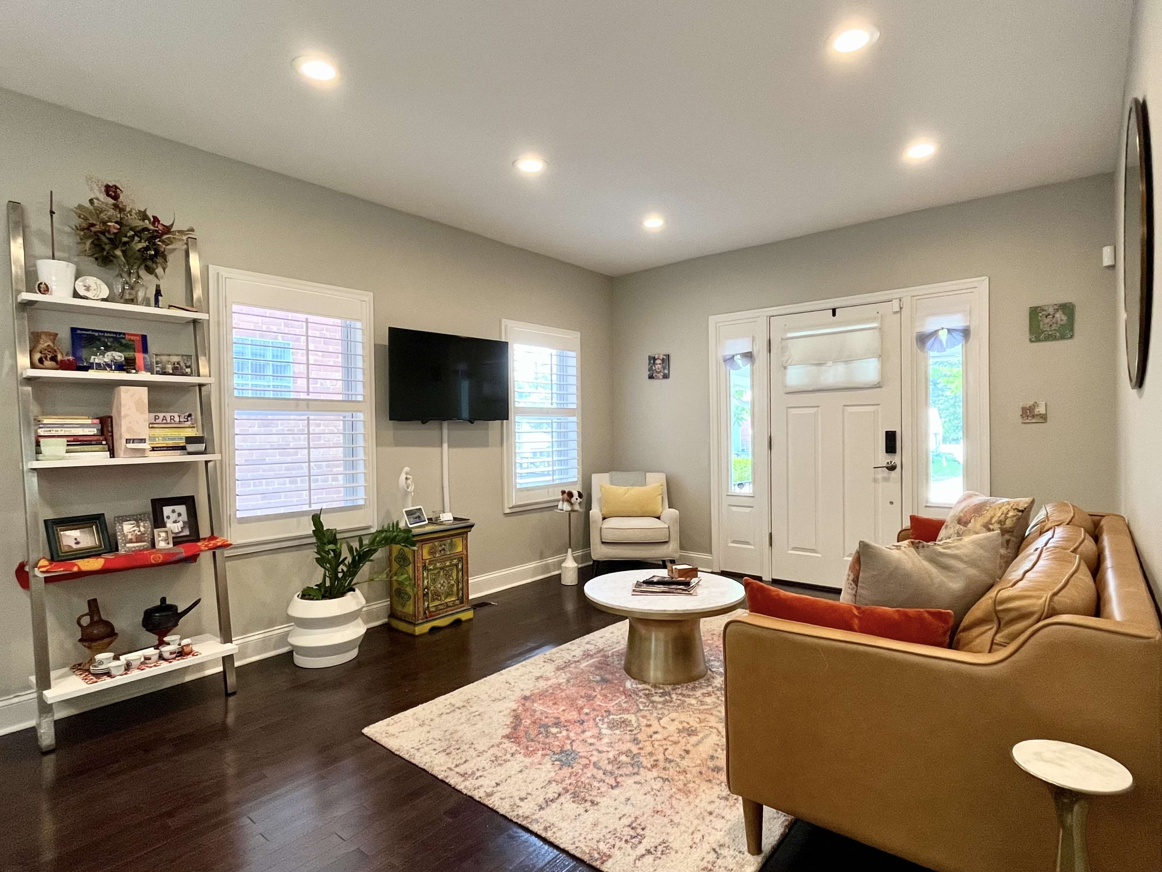 The living room features a brown couch, a round coffee table, and a television mounted on the wall, with a front door and windows allowing natural light.