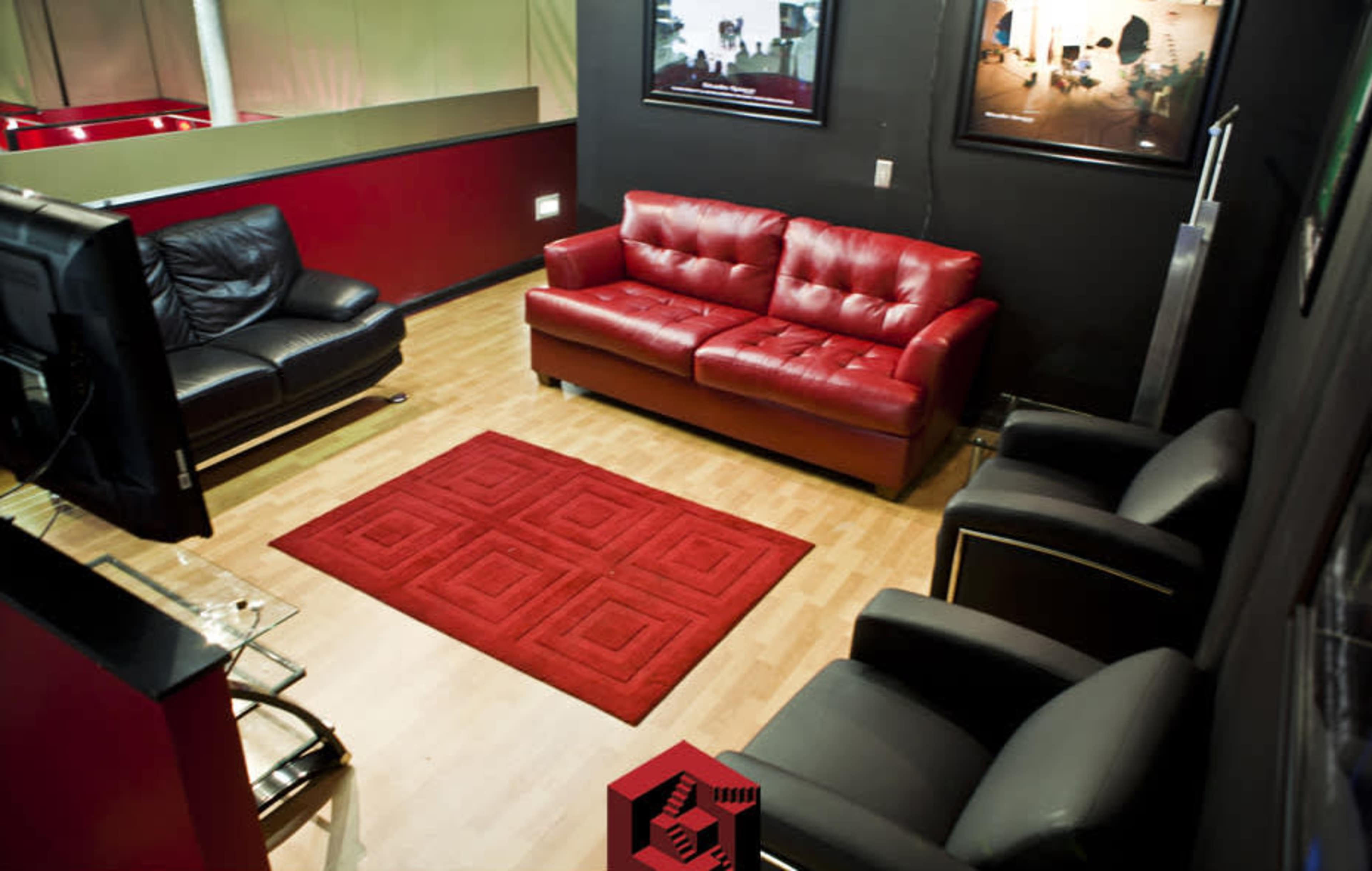 A seating area featuring a black leather couch, a red leather sofa, and two black armchairs on a wooden floor with a red area rug in the center.