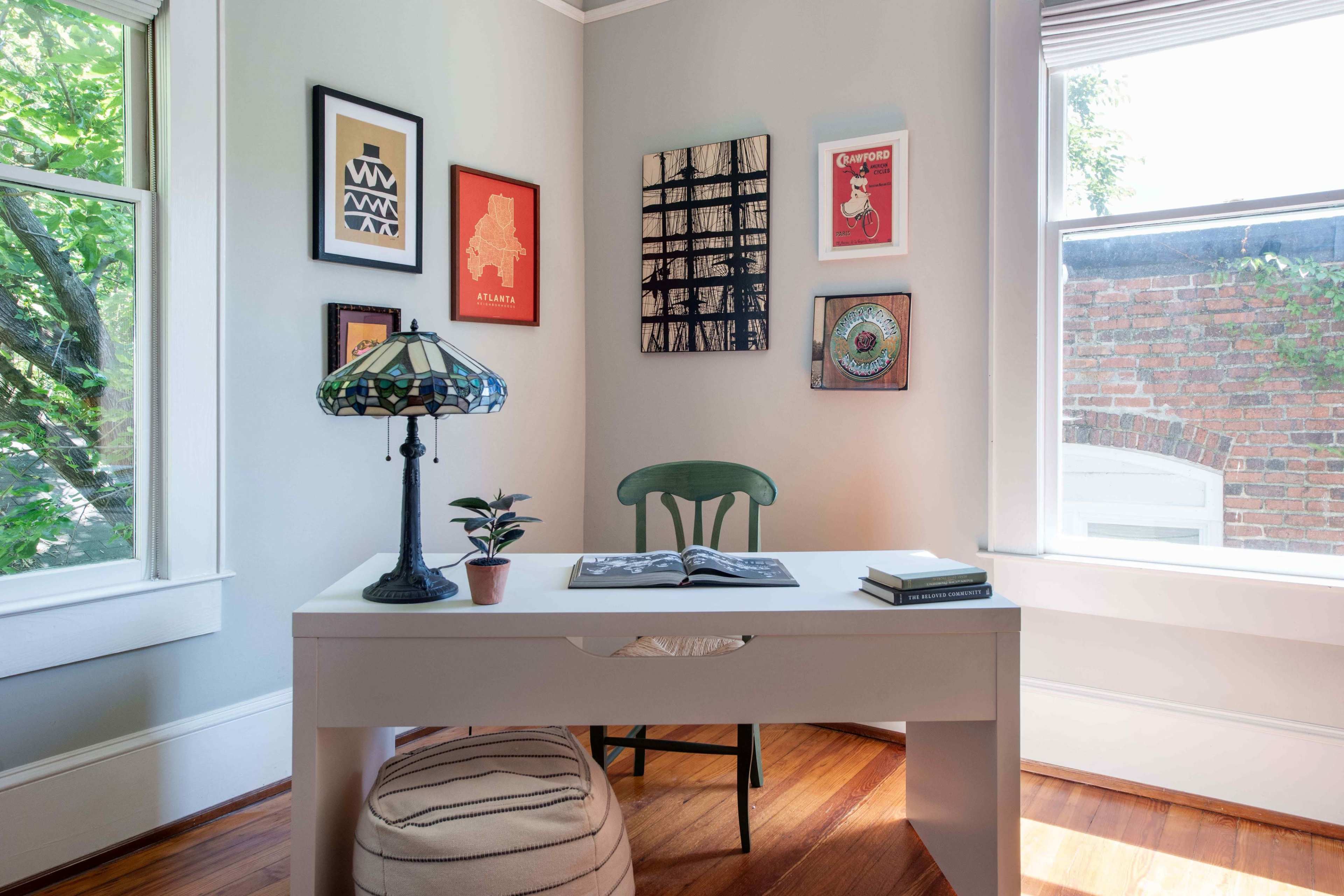 The image shows a small home office with a white desk, a green chair, decorative wall art, and a lamp, all highlighted by natural light from a window.